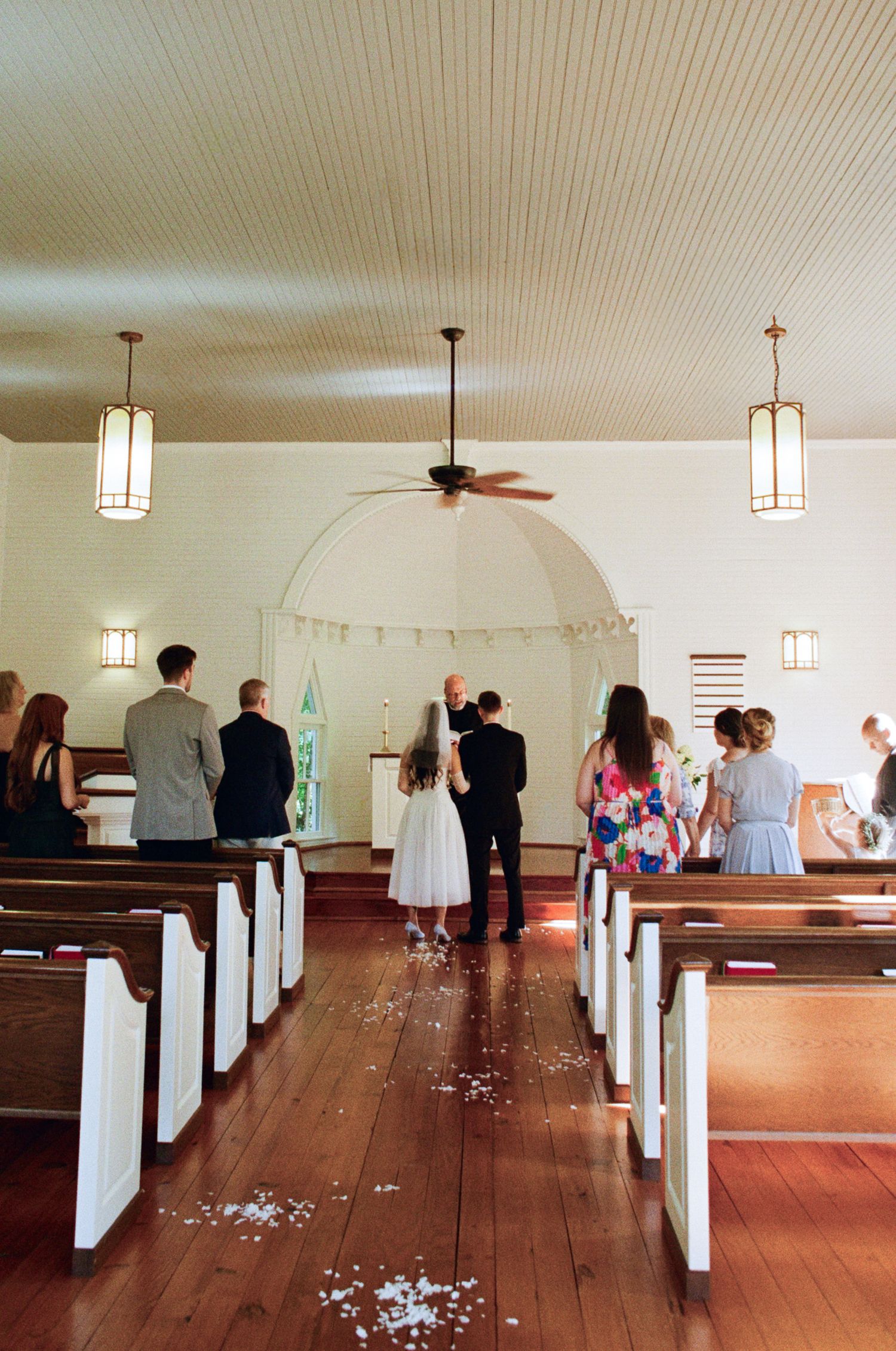 Bride and groom stand before the officiant at the altar of a white wooden chapel as wedding guests watch from the pews.