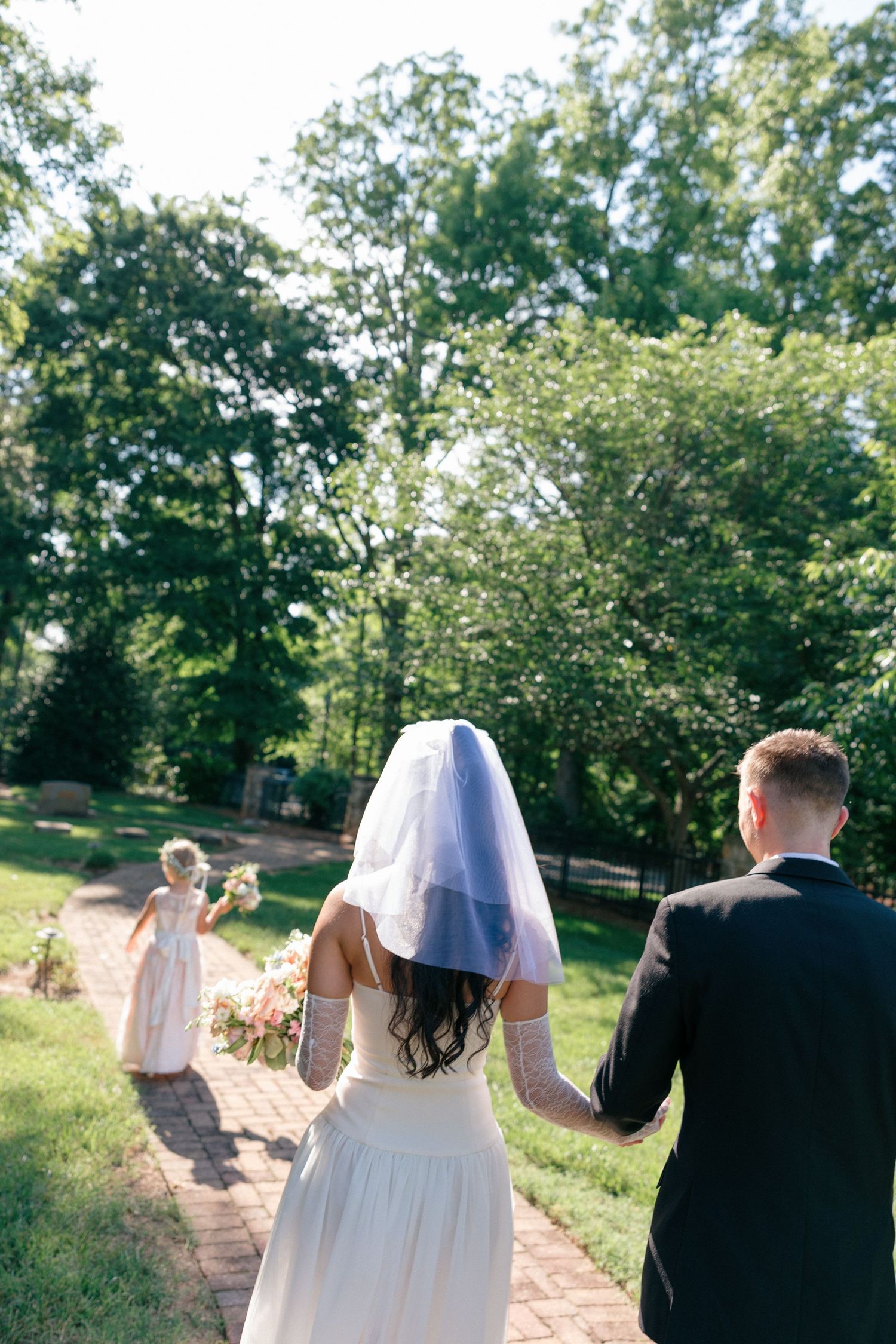 Bride in a short veil and groom in a dark suit walk hand in hand down a brick garden path toward a flower girl surrounded by trees.