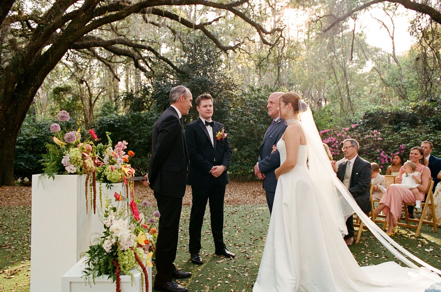 Outdoor garden wedding ceremony with bride in white gown, groom in navy suit, officiant, and floral arrangements under oak trees.