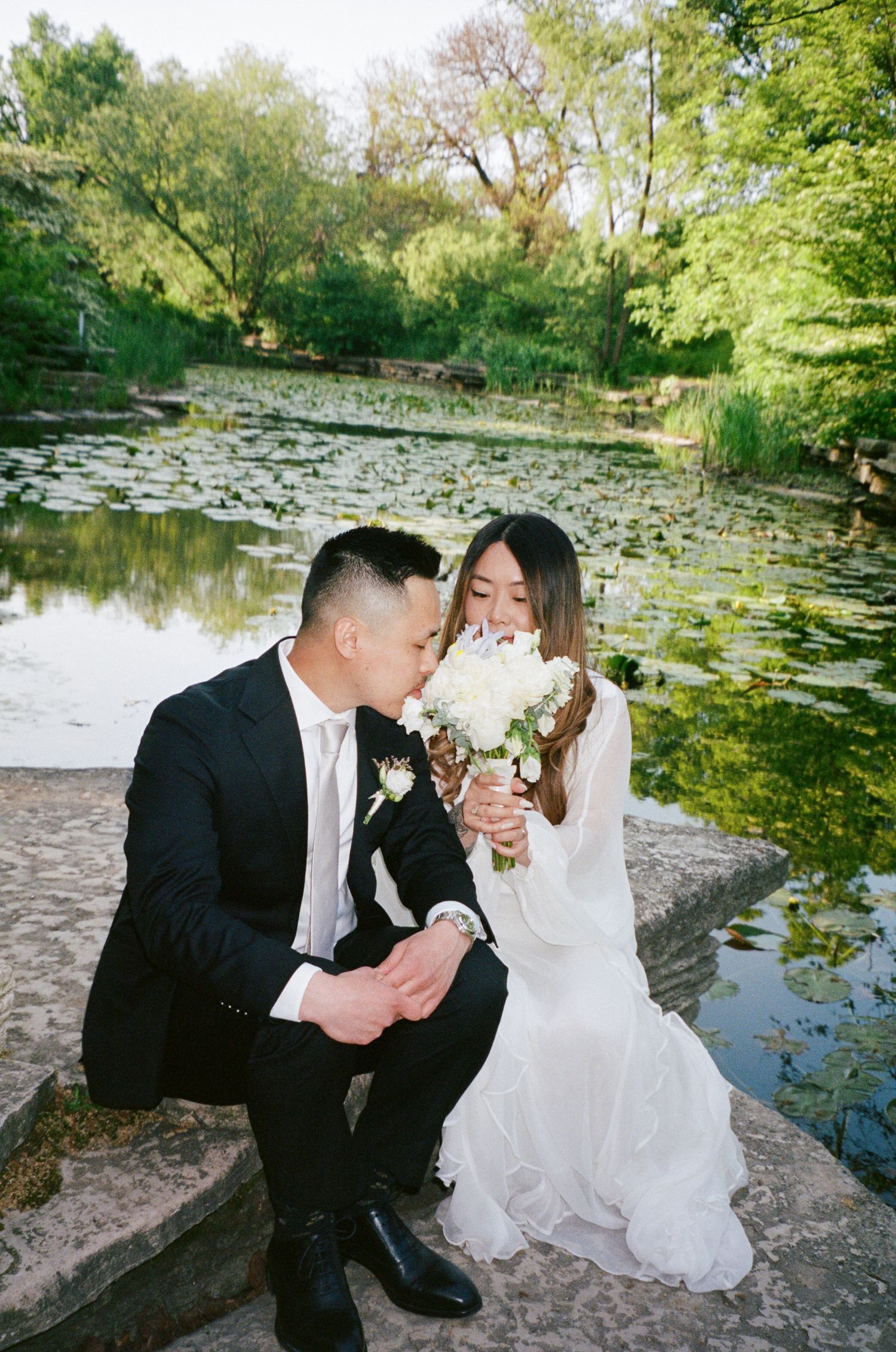 Romantic wedding couple shares tender moments by a serene lily pond in a lush garden setting.