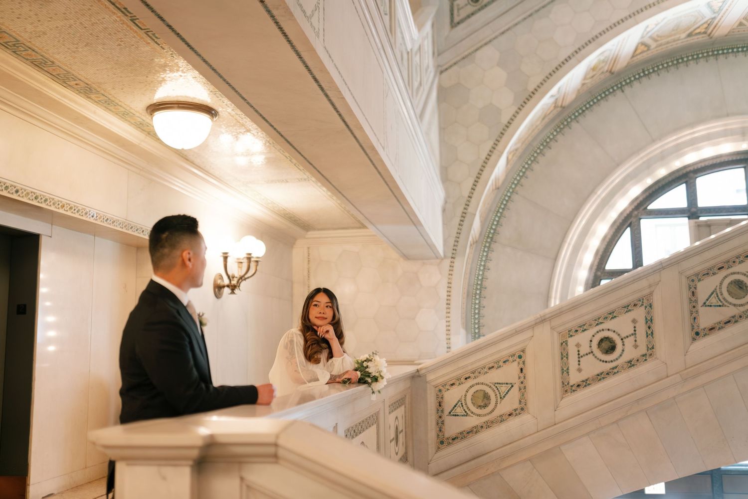 Two people converse on an elegant white marble staircase with ornate details and arched architecture.