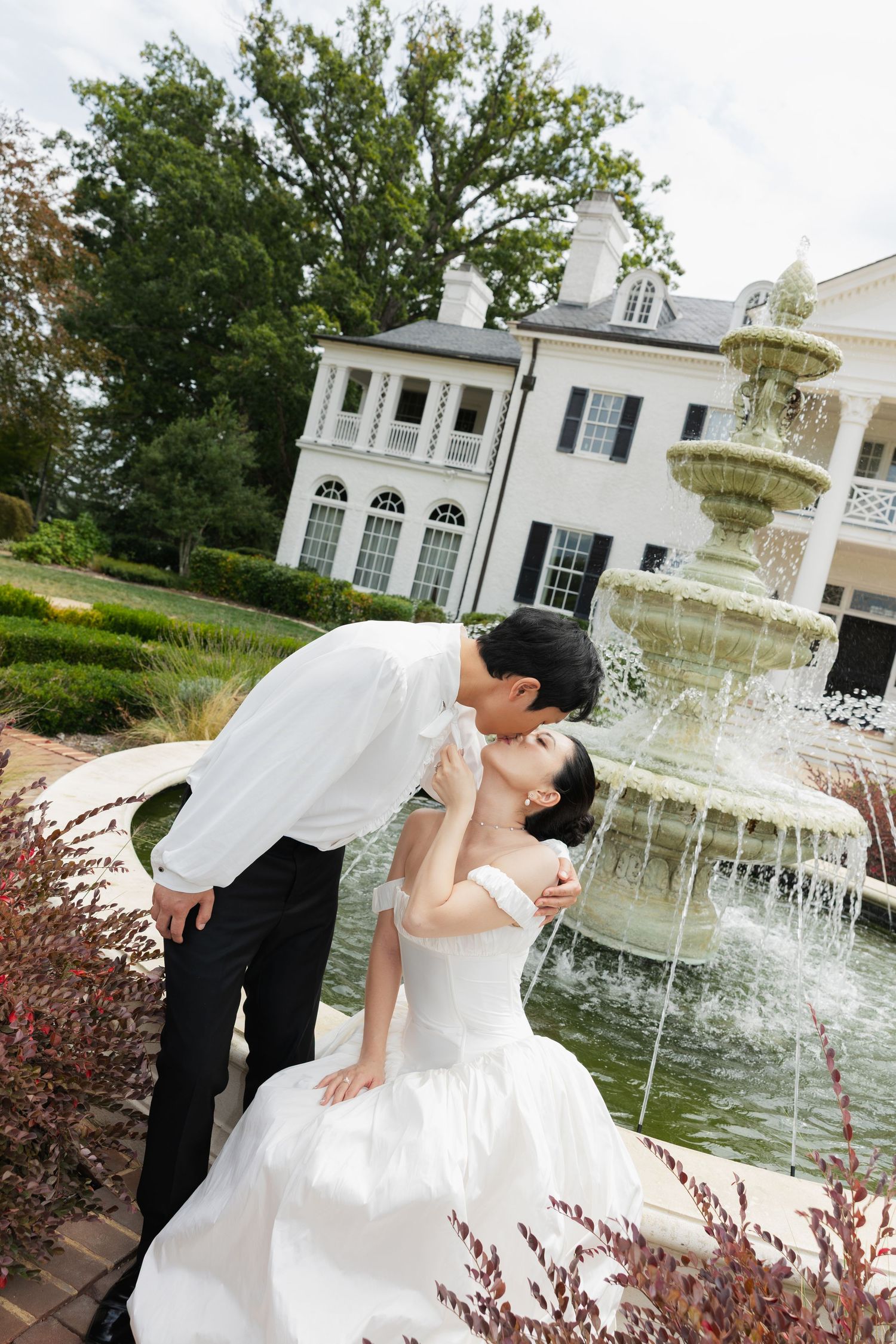 A romantic wedding couple dips and poses for photos in front of an elegant white mansion with a fountain in the background.