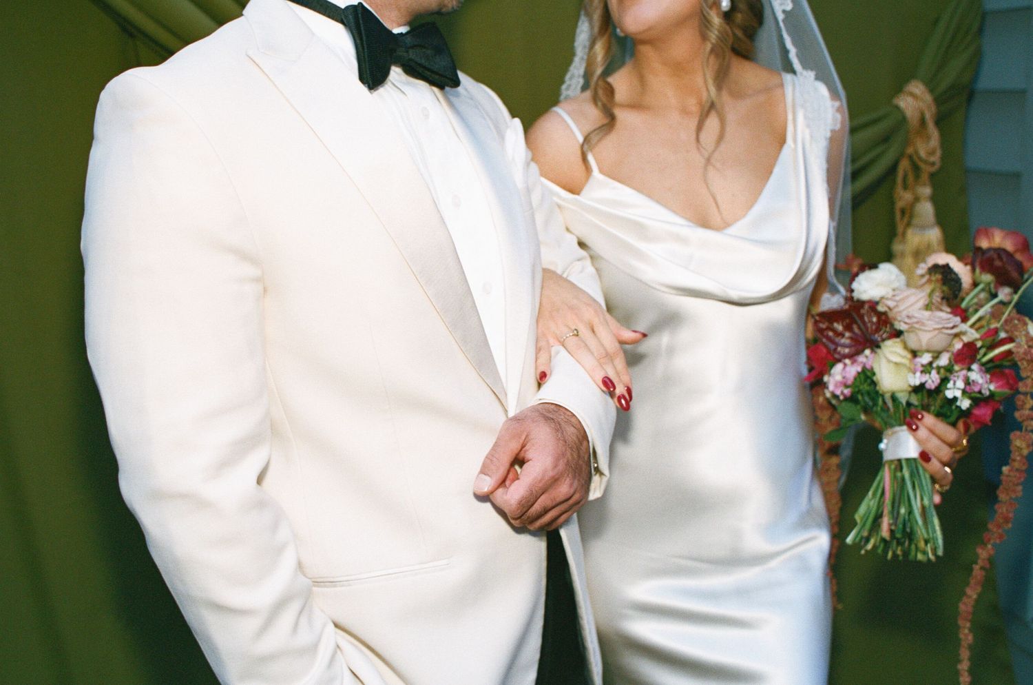 Elegant bride and groom in white satin gown and ivory tuxedo holding hands at wedding ceremony.