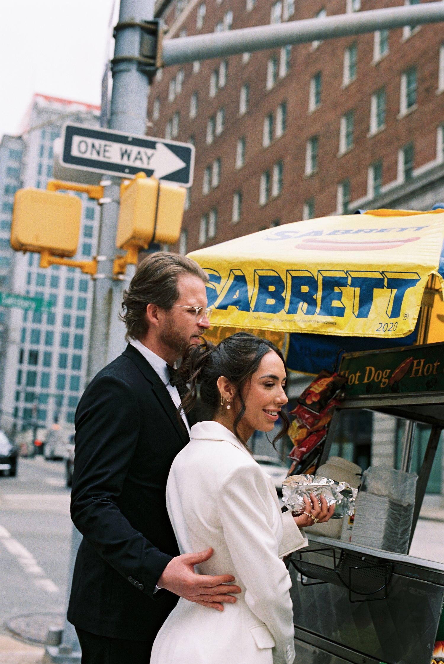 Bride and groom embrace at a Sabrett hot dog cart on a New York City street corner on their wedding day.