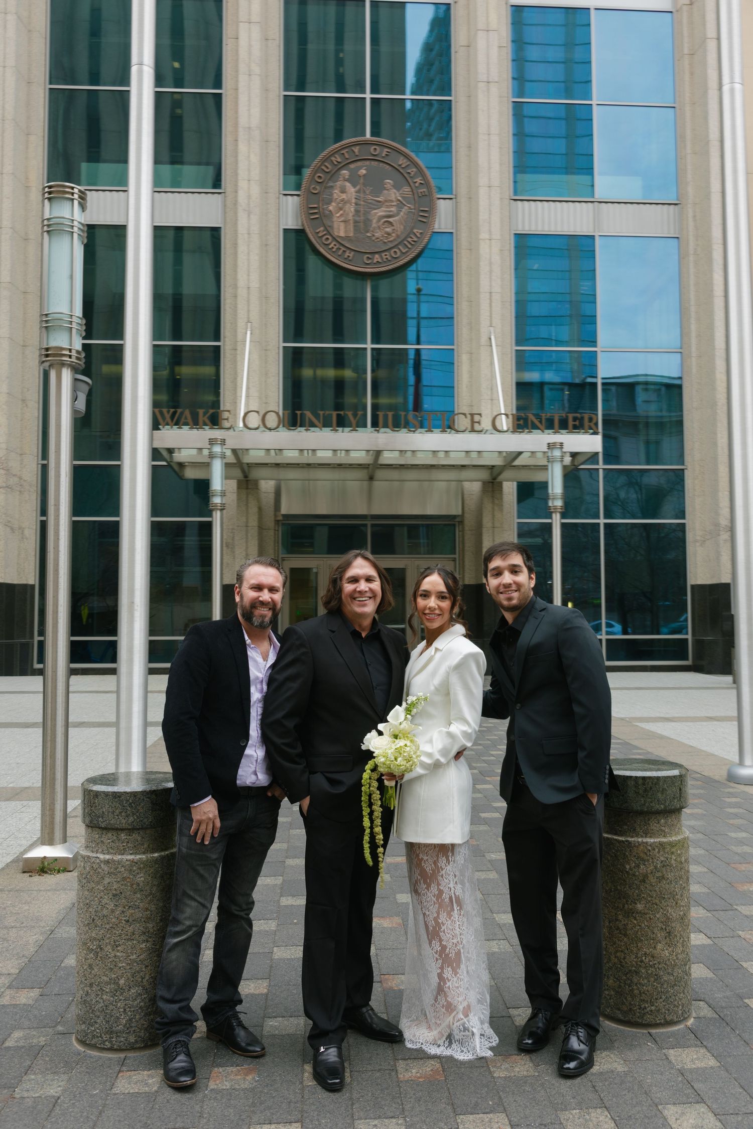 Wedding party of four posing outside Wake County Justice Center, bride in white suit holding bouquet.