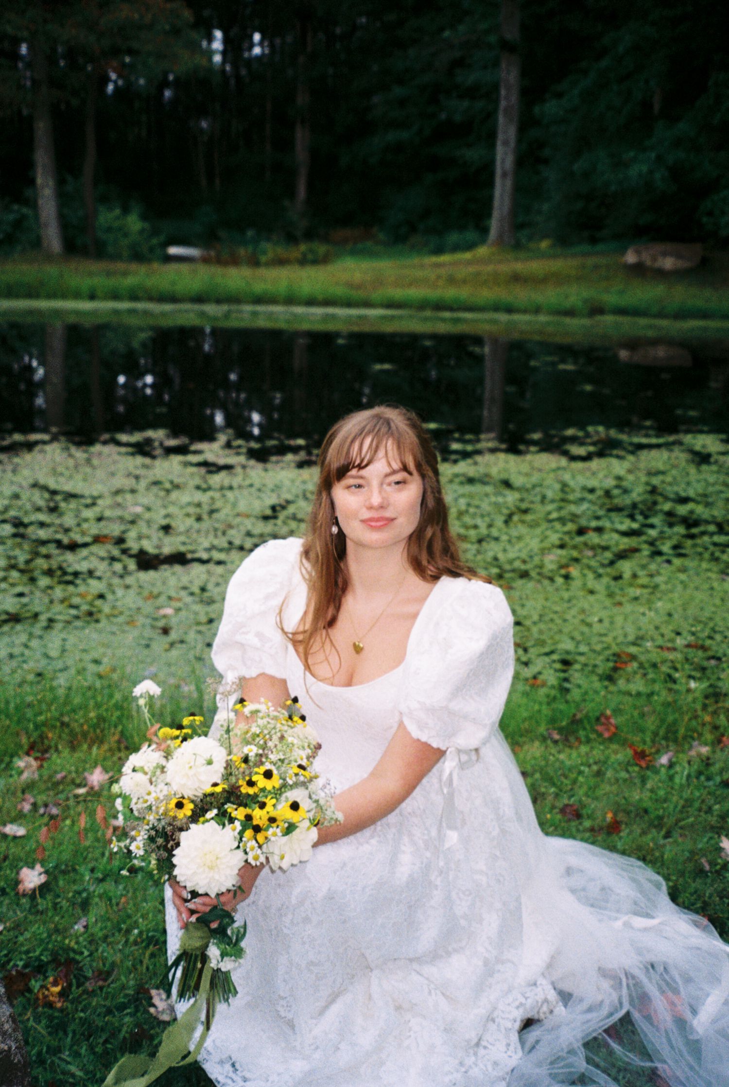 A person in a white dress poses gracefully by a serene pond with lily pads and flowers in a garden setting.