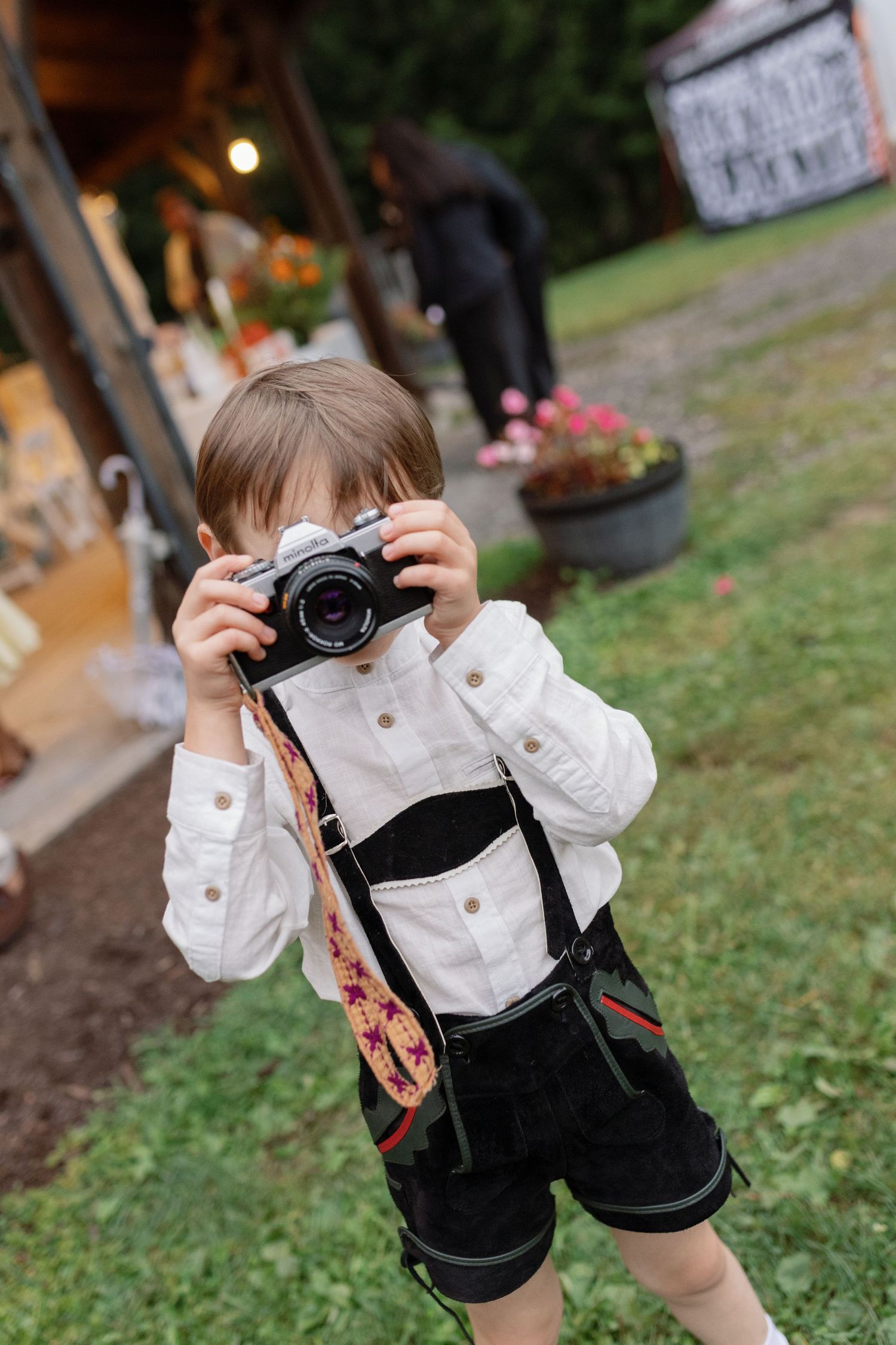 A young photographer in black overalls and white shirt takes pictures with a camera in an outdoor garden setting.