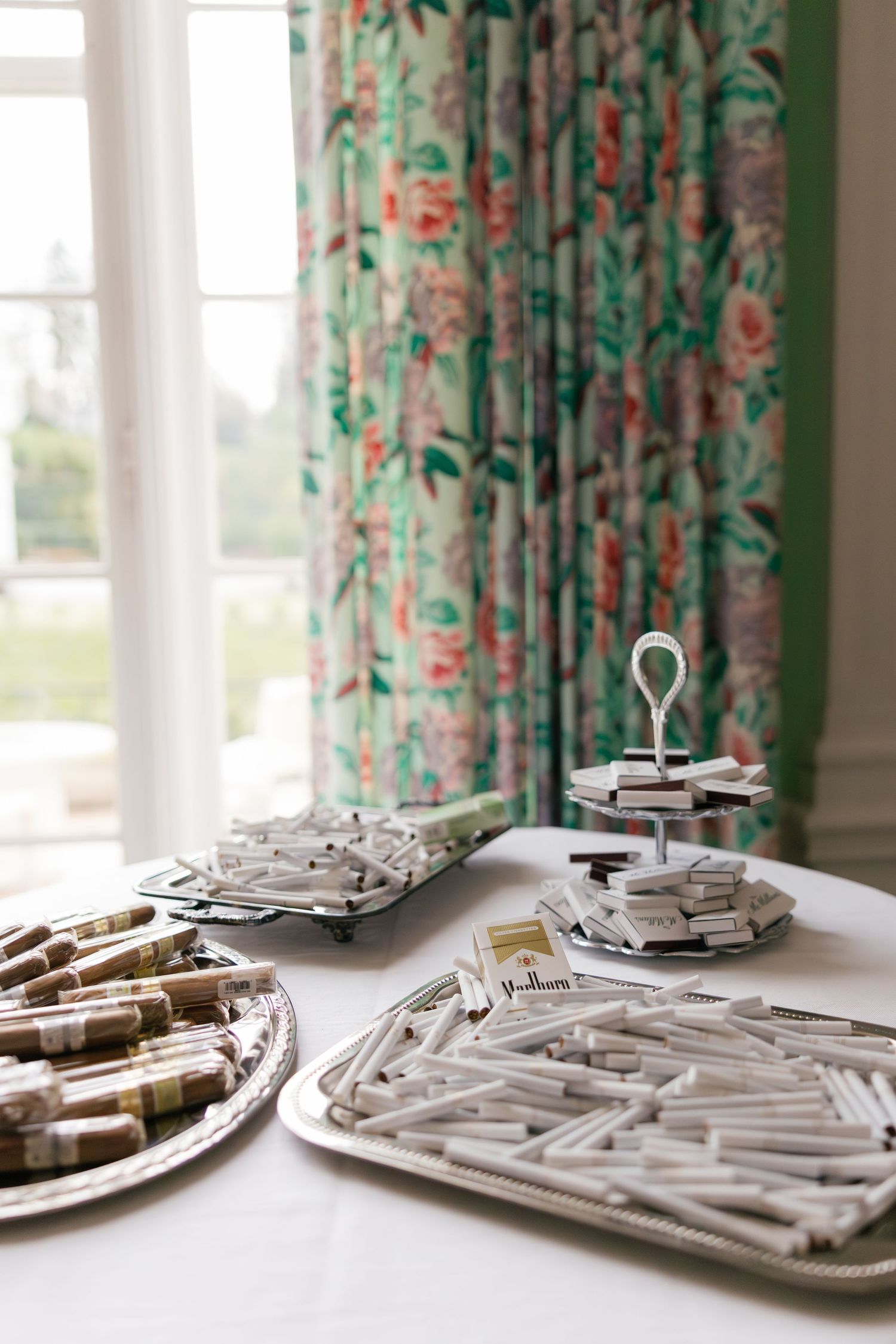 Elegant table display of cigarettes and cigars on silver trays near floral curtains at a formal event.