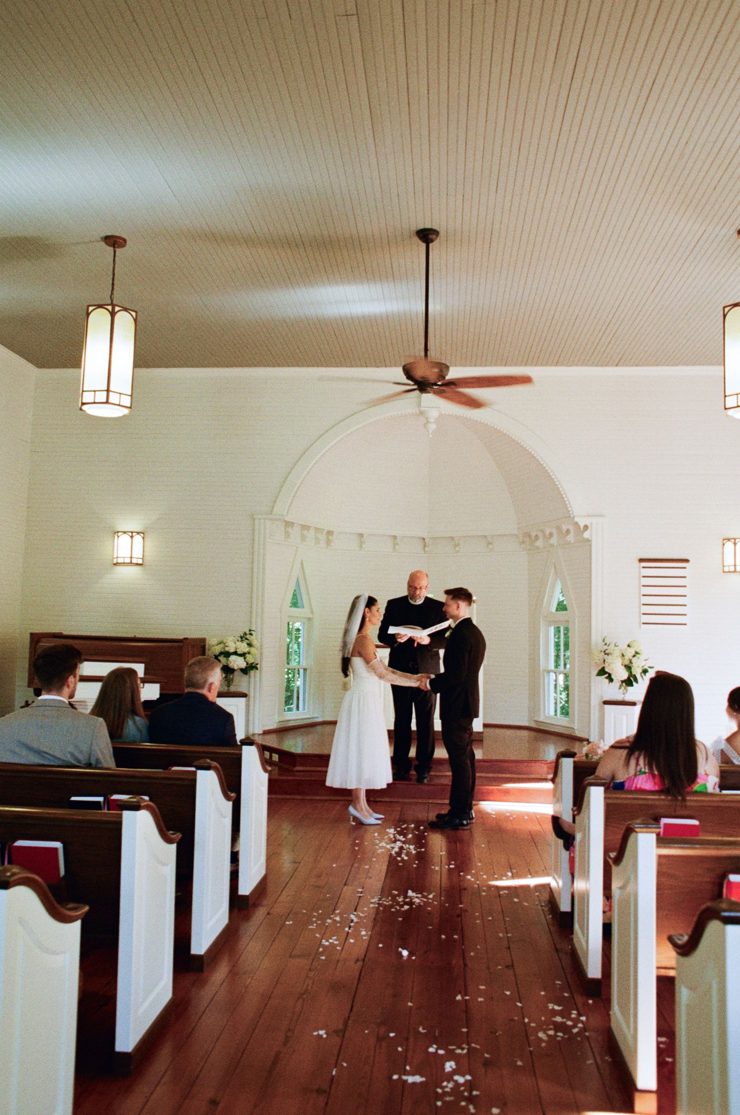 Bride and groom exchange vows before an officiant at the altar of a small white chapel with wooden pews and scattered petals on the aisle.