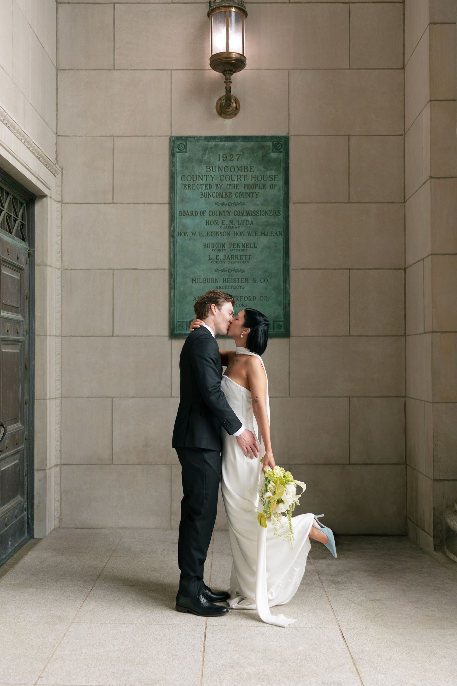 A bride in a white gown and a groom in a dark suit kiss beside a bronze plaque on a stone wall, her pale blue heel lifted behind her.