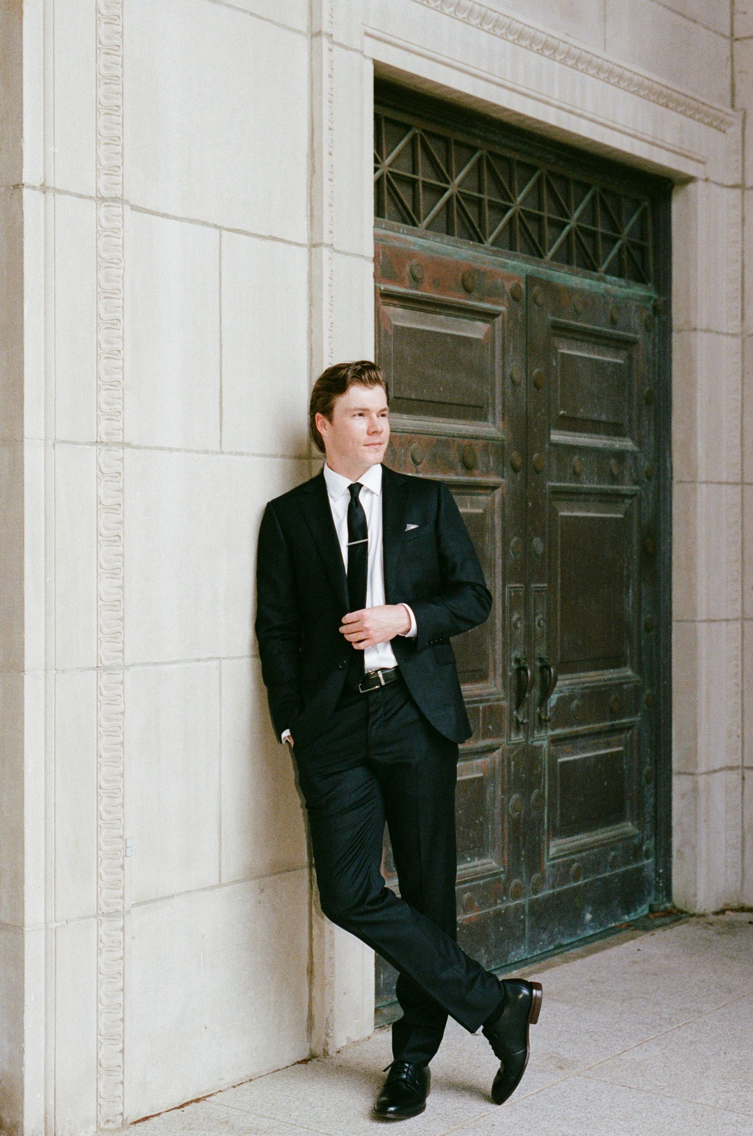 Young man in a black suit and tie leans casually against a limestone wall beside a large ornate bronze door.