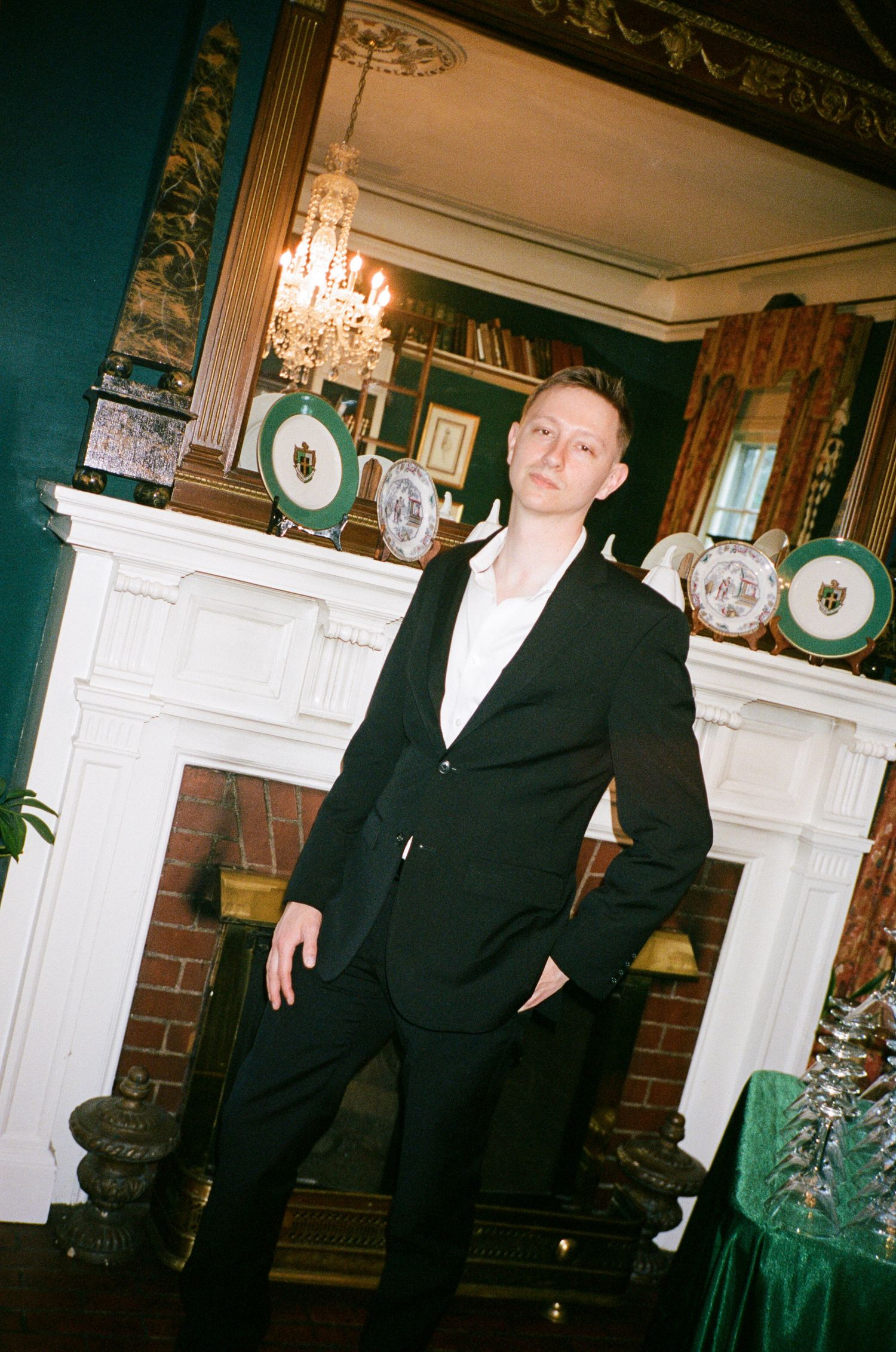 Man in a black blazer and white shirt standing by a white fireplace in an ornate room with a chandelier and decorative plates.