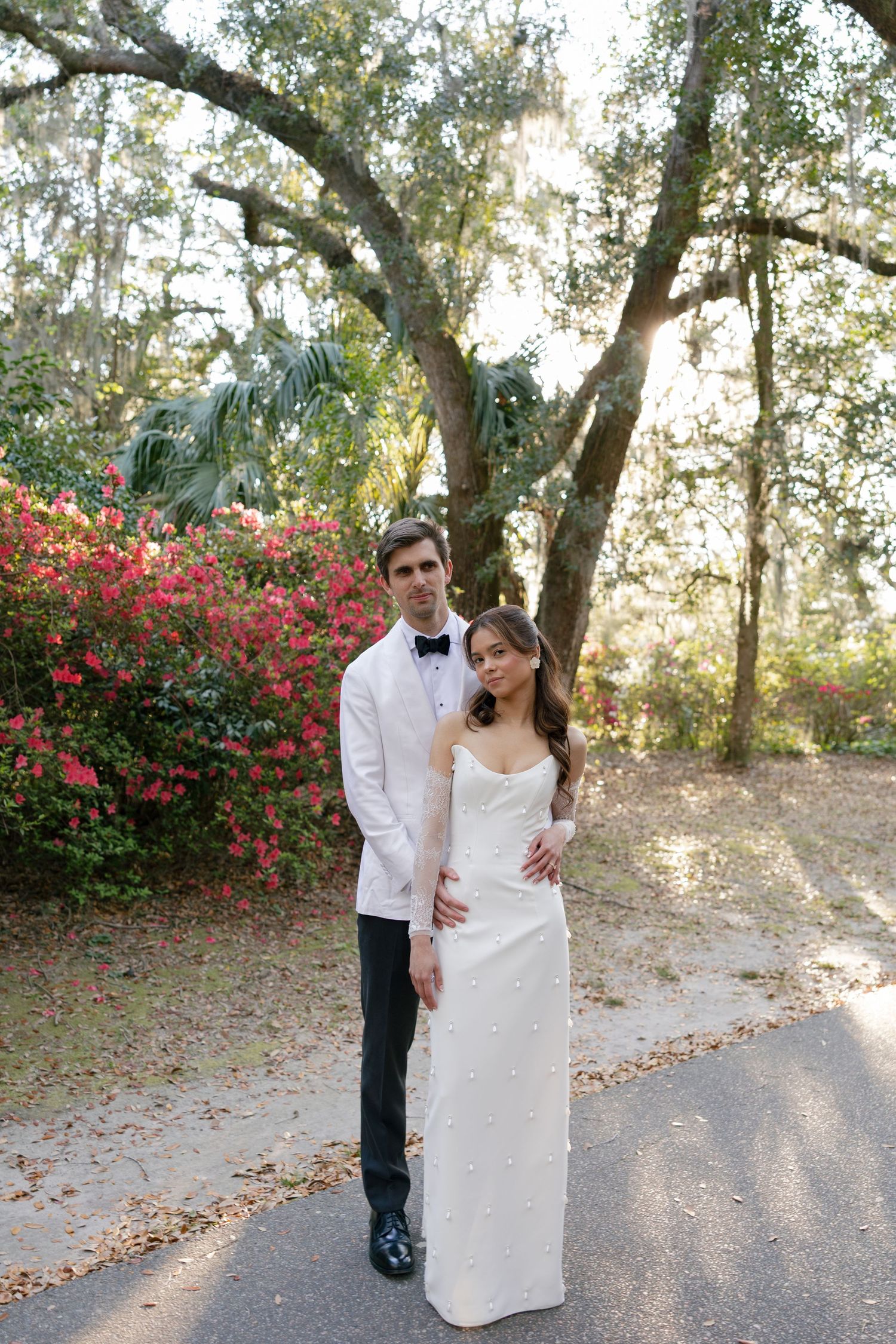 A couple poses together outdoors on a sun-dappled path lined with blooming azalea bushes and oak trees.