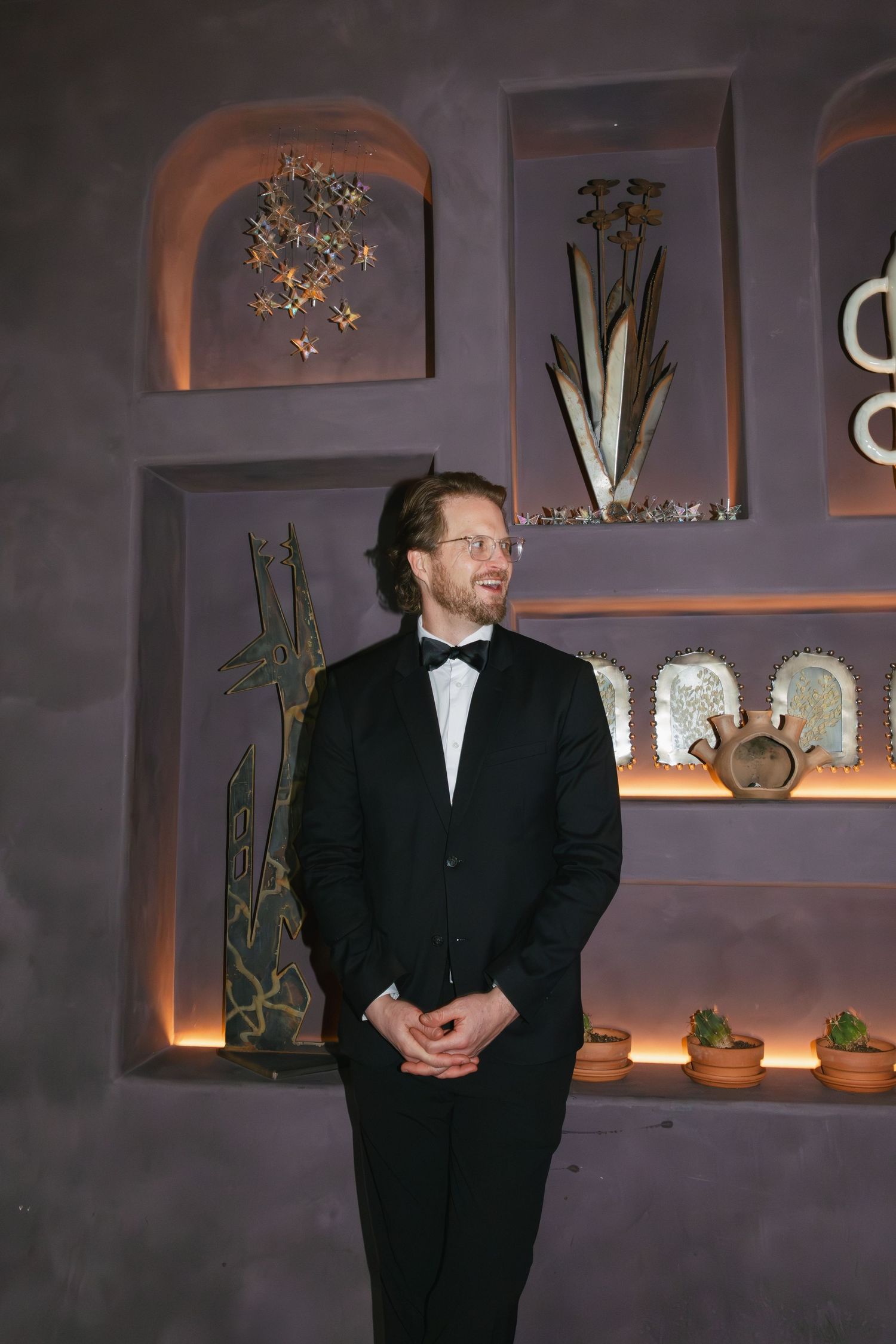 Man in black tuxedo and bow tie standing near decorative wall niches with art and plants.