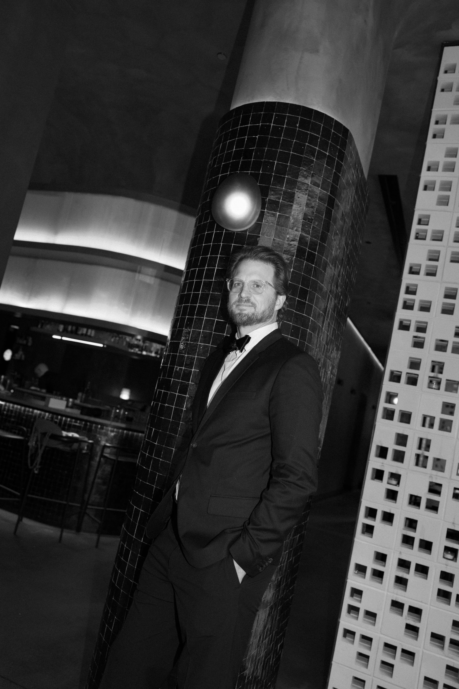 Black and white photo of a man in a tuxedo leaning against a tiled column in an elegant venue.