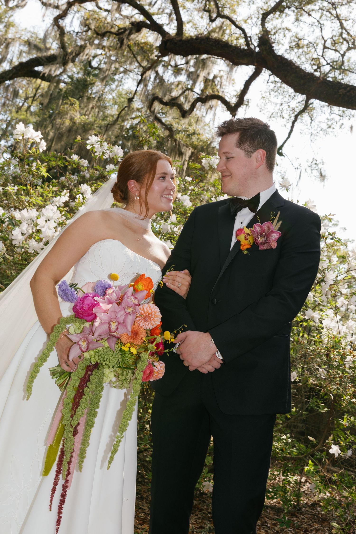Bride and groom smile at each other outdoors, she holds a colorful bouquet, he wears a black tuxedo with floral boutonniere.