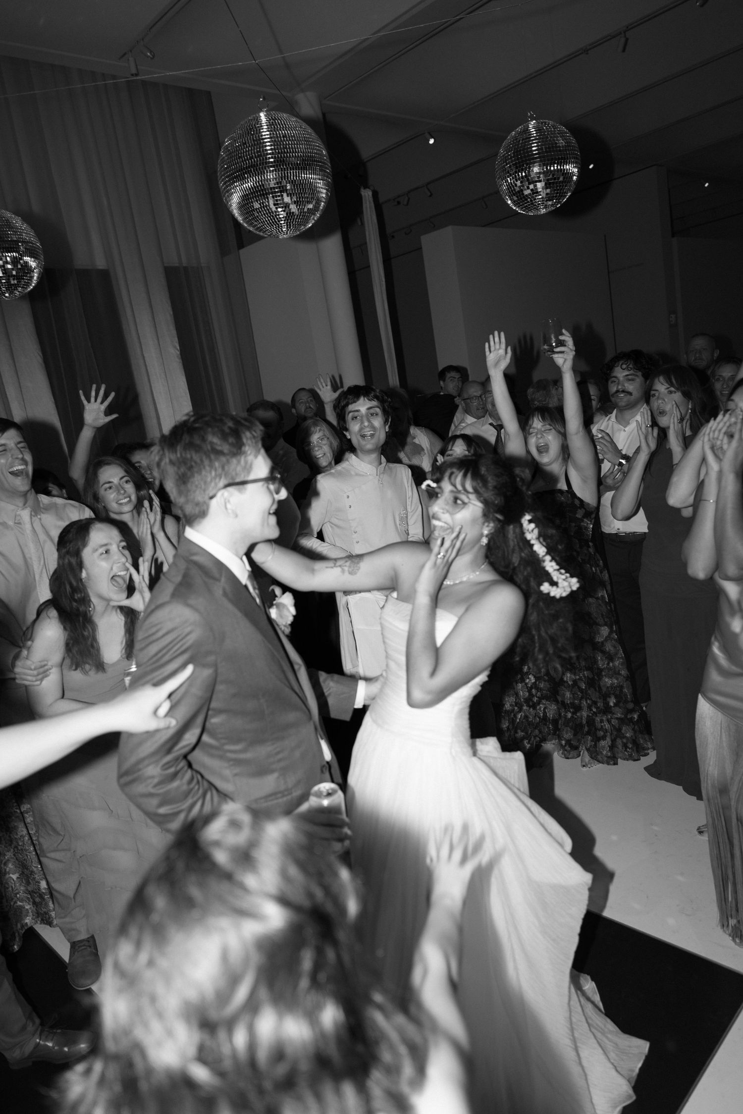 Black and white photo of bride and groom dancing joyfully at wedding reception with excited guests and disco balls.