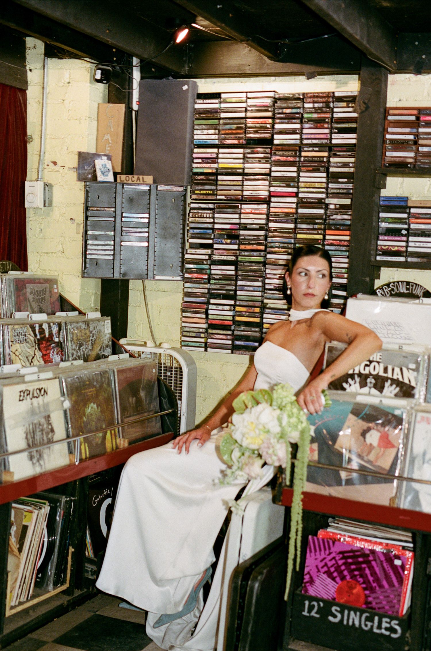 Bride in a white strapless gown holding a white floral bouquet sits among vinyl records and cassette tapes in a vintage record store.