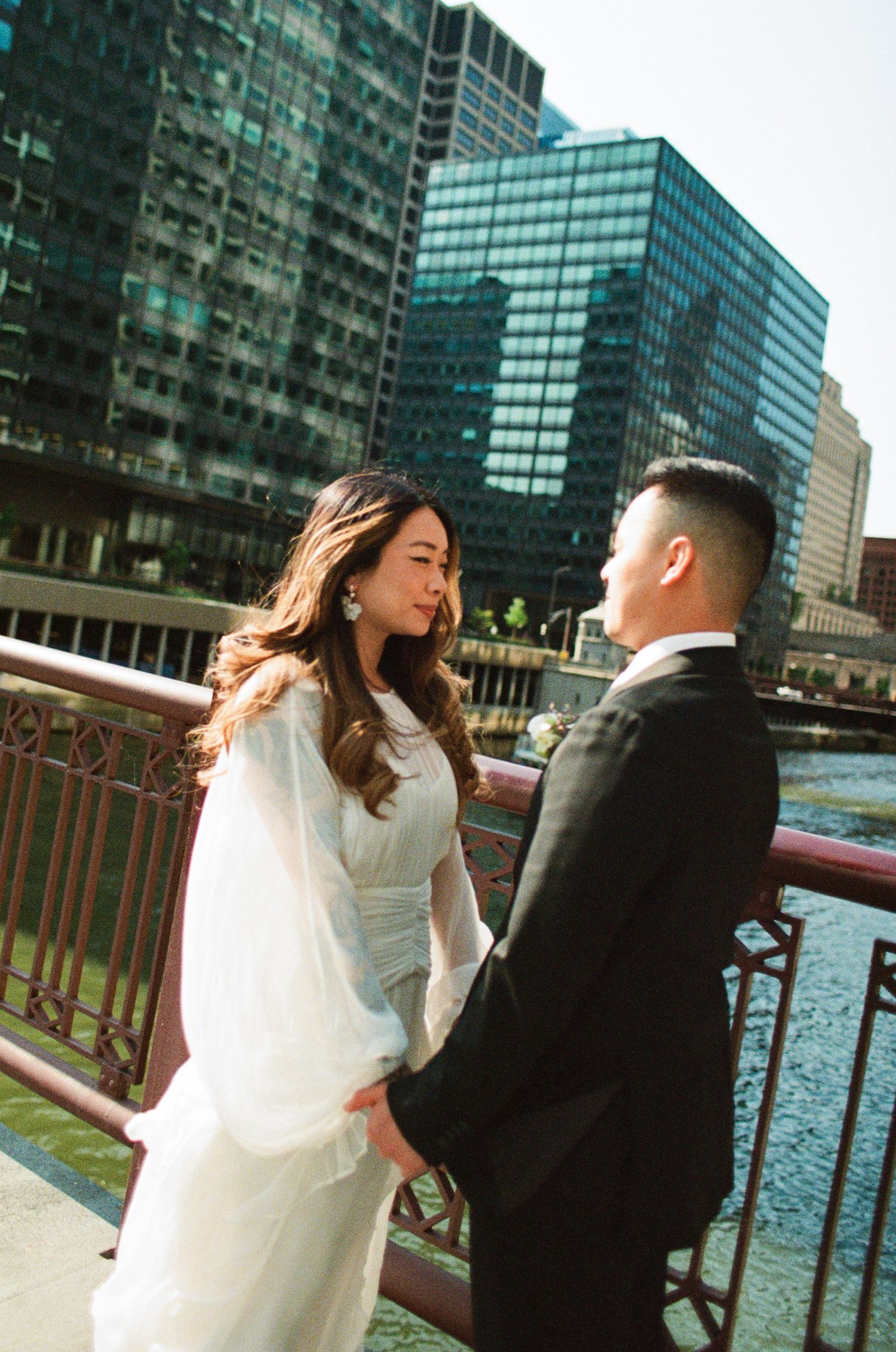 Romantic couple in wedding attire share candid moments on a bridge with modern glass buildings in the background.