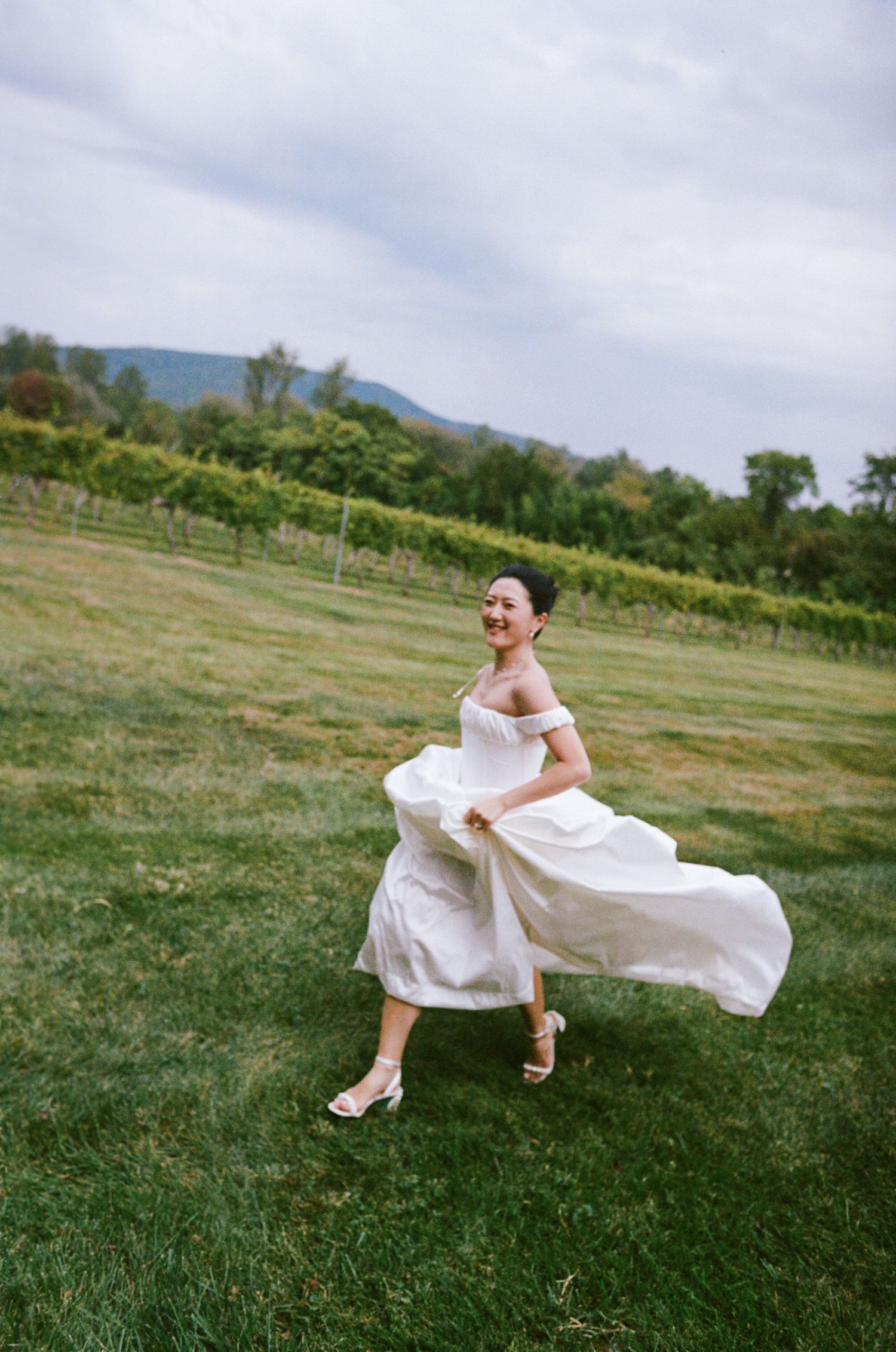 A figure in a flowing white dress dances and twirls gracefully across a green hillside meadow on a cloudy summer day.