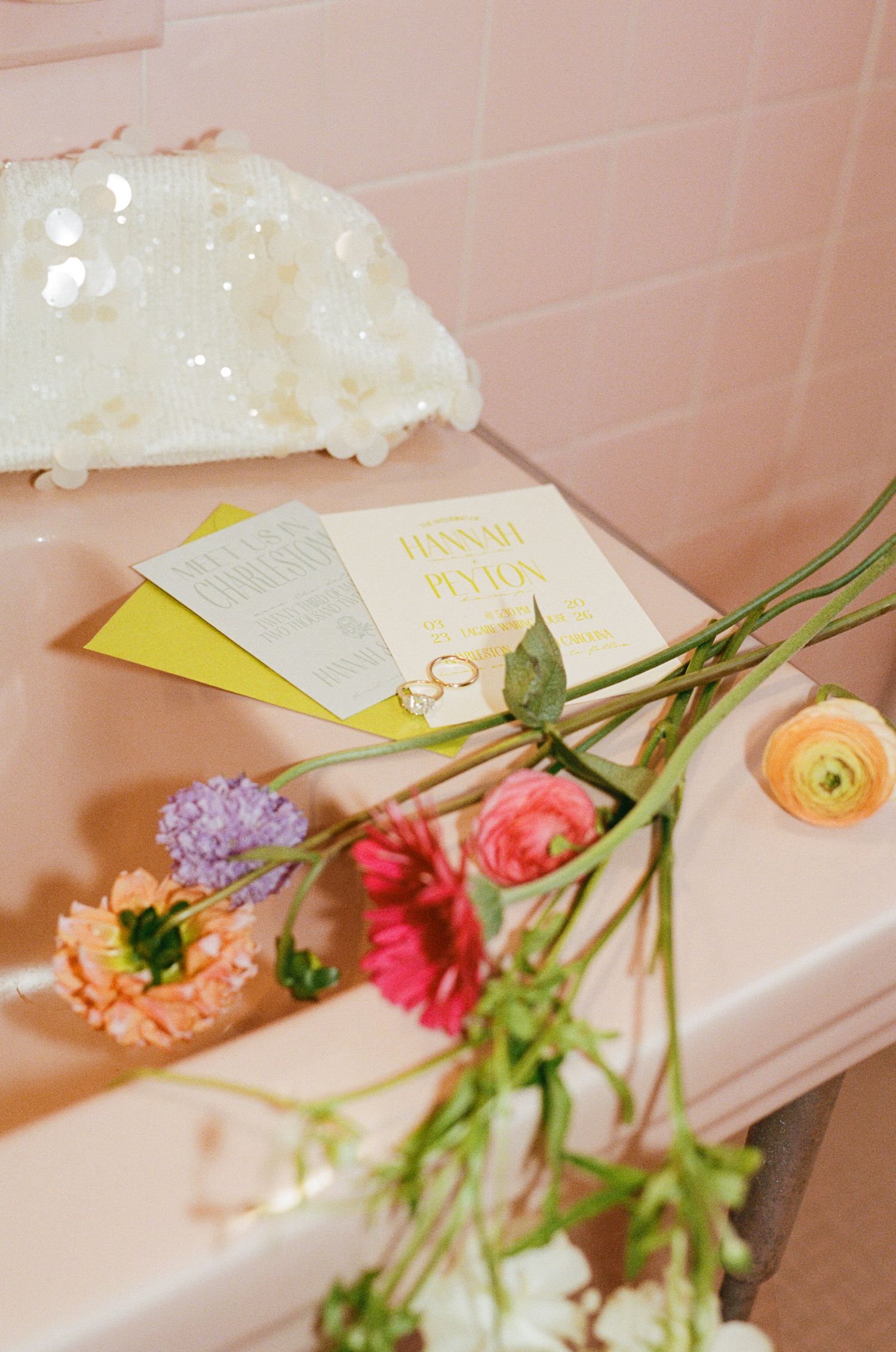 Pink bathtub with colorful flowers, wedding rings, and yellow invitation cards in a romantic bridal flat lay.