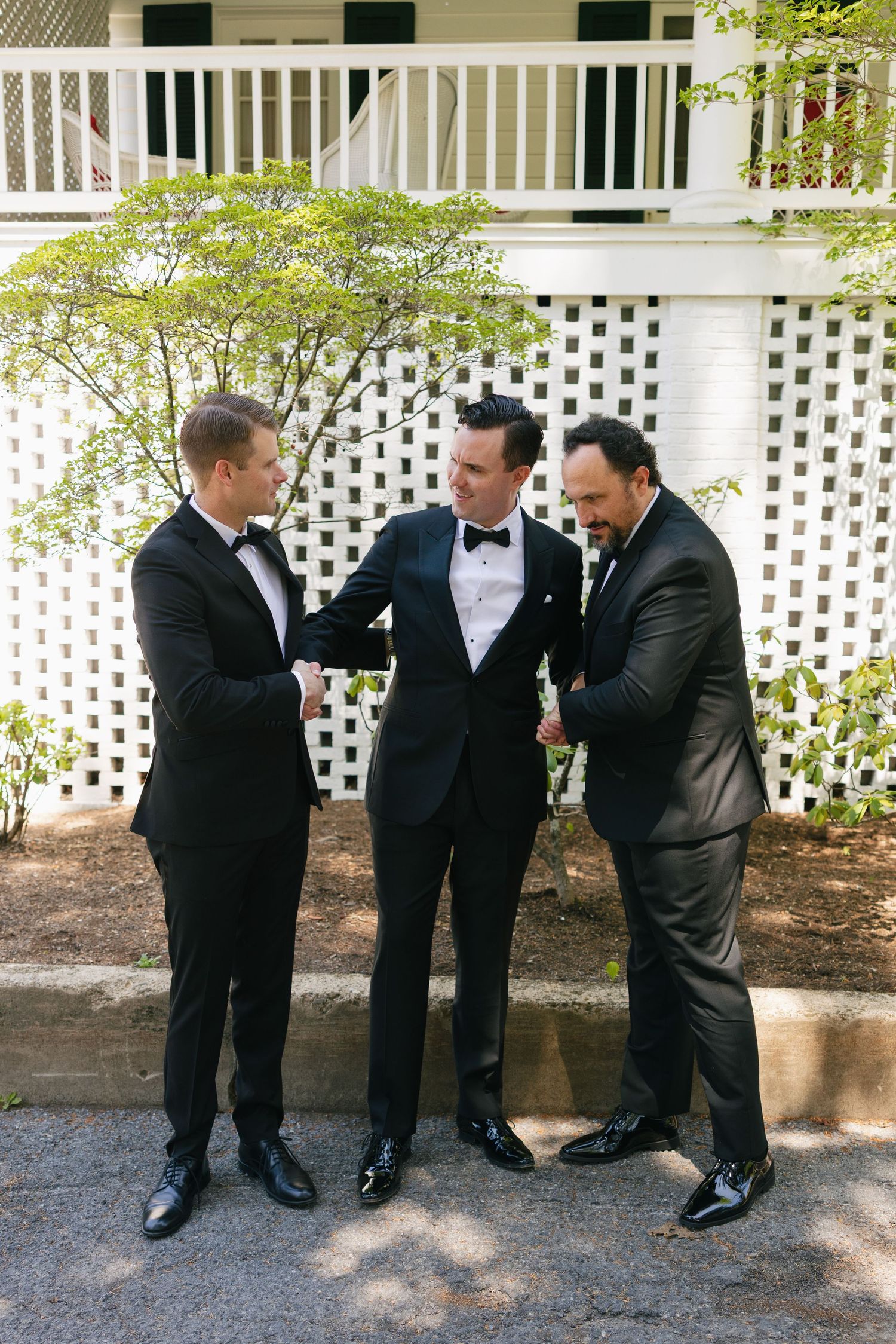 Three men in black tuxedos sharing a celebratory moment outdoors at a wedding venue.