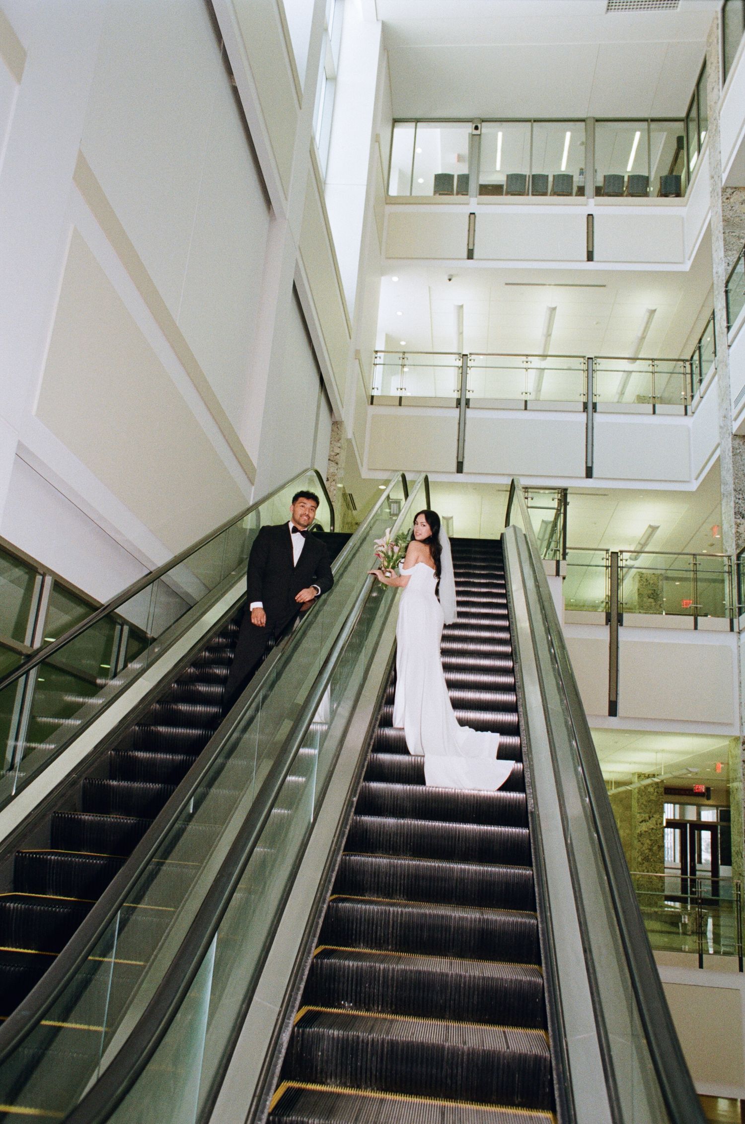 A couple in formal wedding attire stands on opposite sides of an escalator in a modern building atrium.