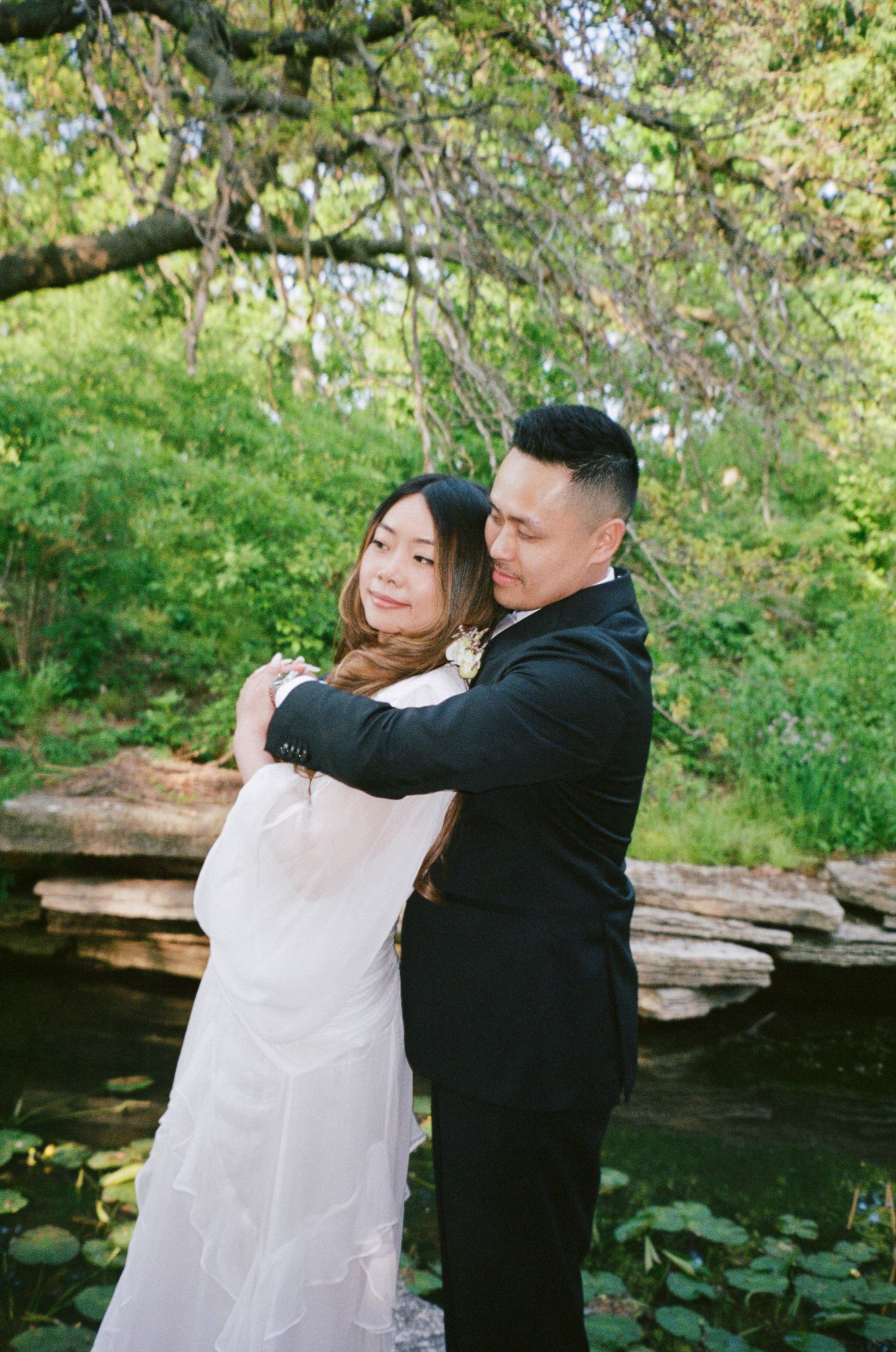 A couple embraces in a romantic moment by a pond surrounded by lush greenery and blooming trees in a garden setting.