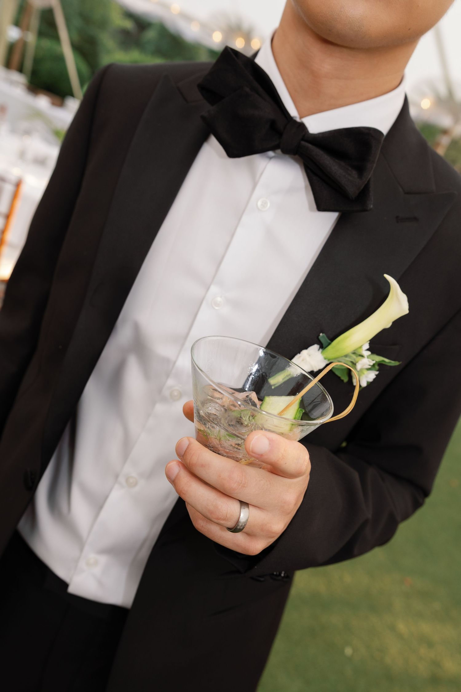 A close-up of a groom in black tuxedo and bowtie holding a cocktail glass with a calla lily boutonniere.