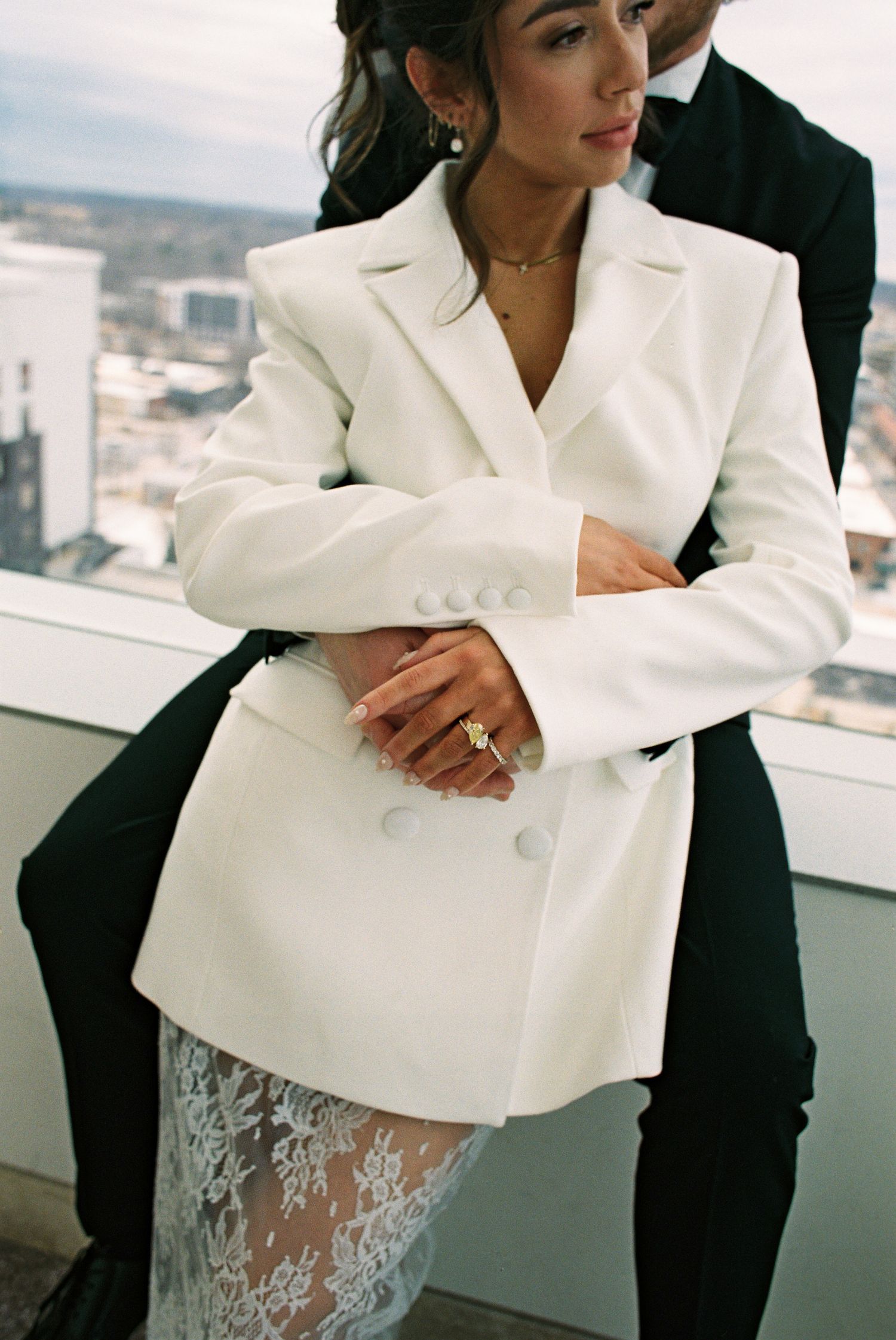 Bride in white double-breasted blazer and lace skirt embraced by groom in black suit on rooftop terrace.