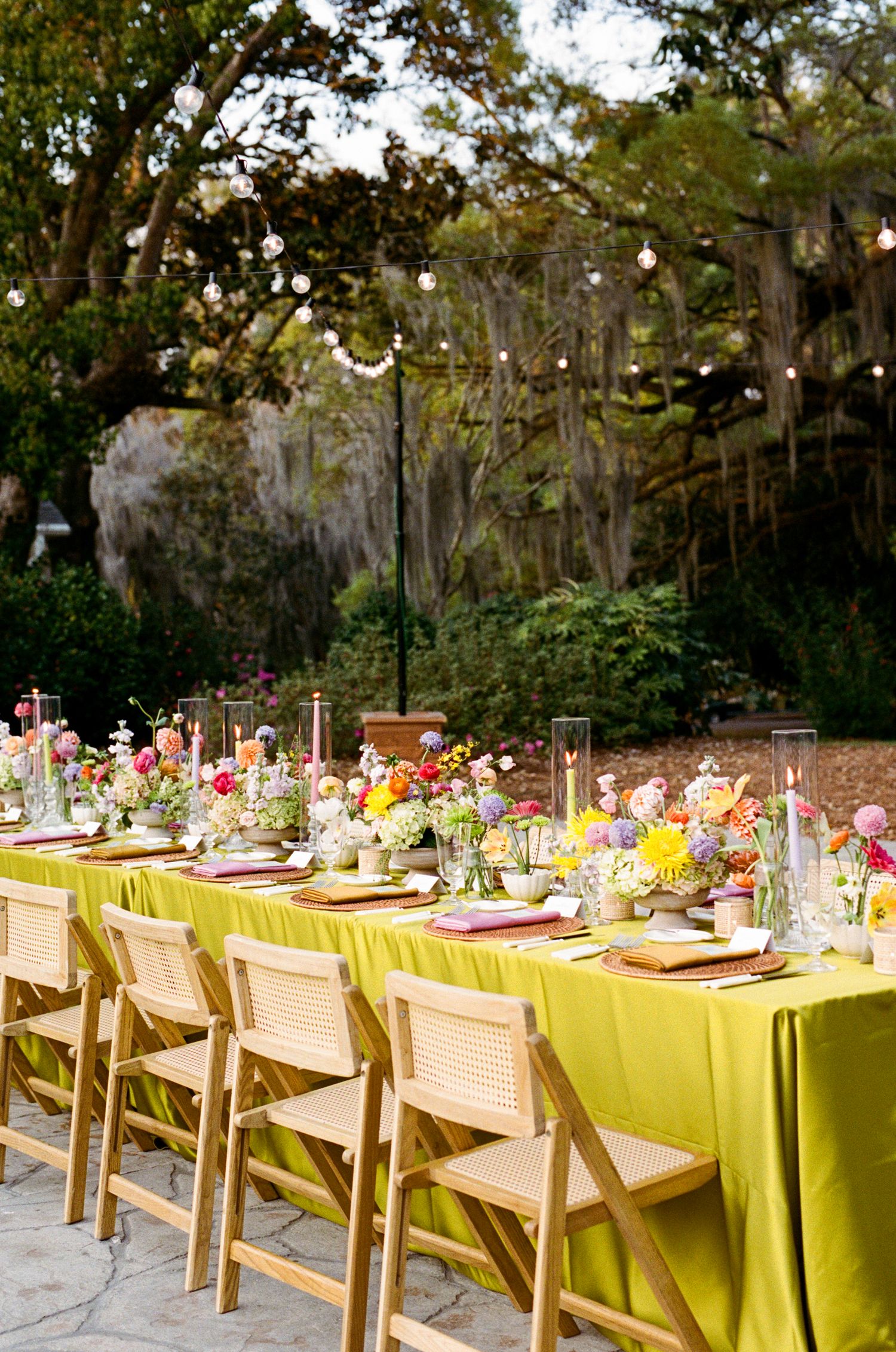 Outdoor wedding reception with chartreuse tablecloth, colorful floral centerpieces, wooden chairs, and string lights among trees.