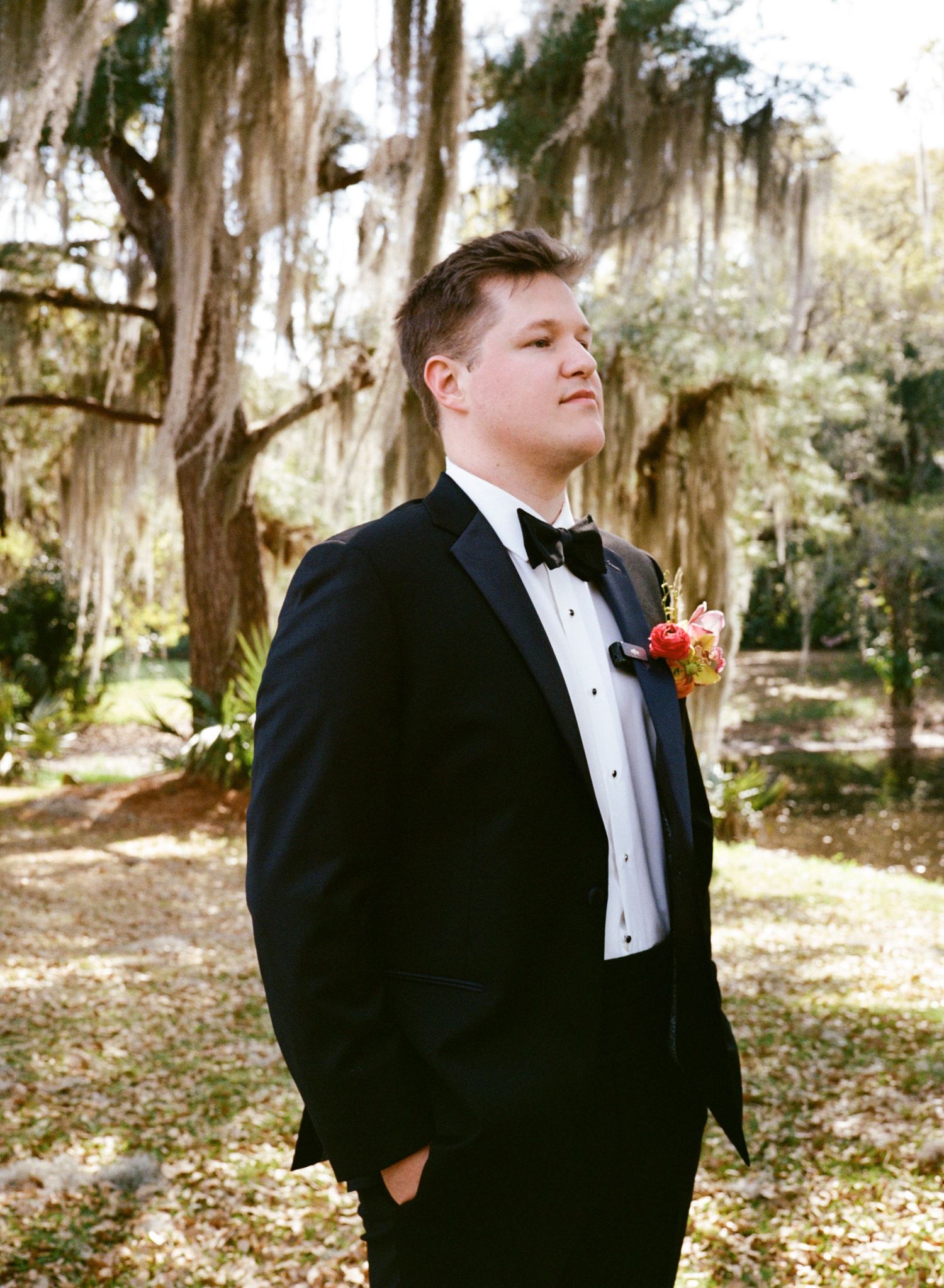 Groom in black tuxedo with bow tie and boutonniere standing outdoors under Spanish moss-draped oak trees.