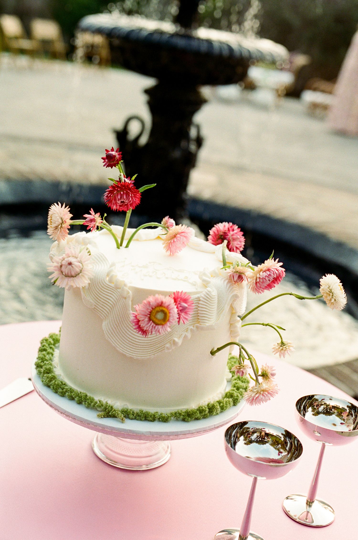 White wedding cake with pink daisy flowers on a pedestal stand, displayed outdoors near a fountain with silver goblets.