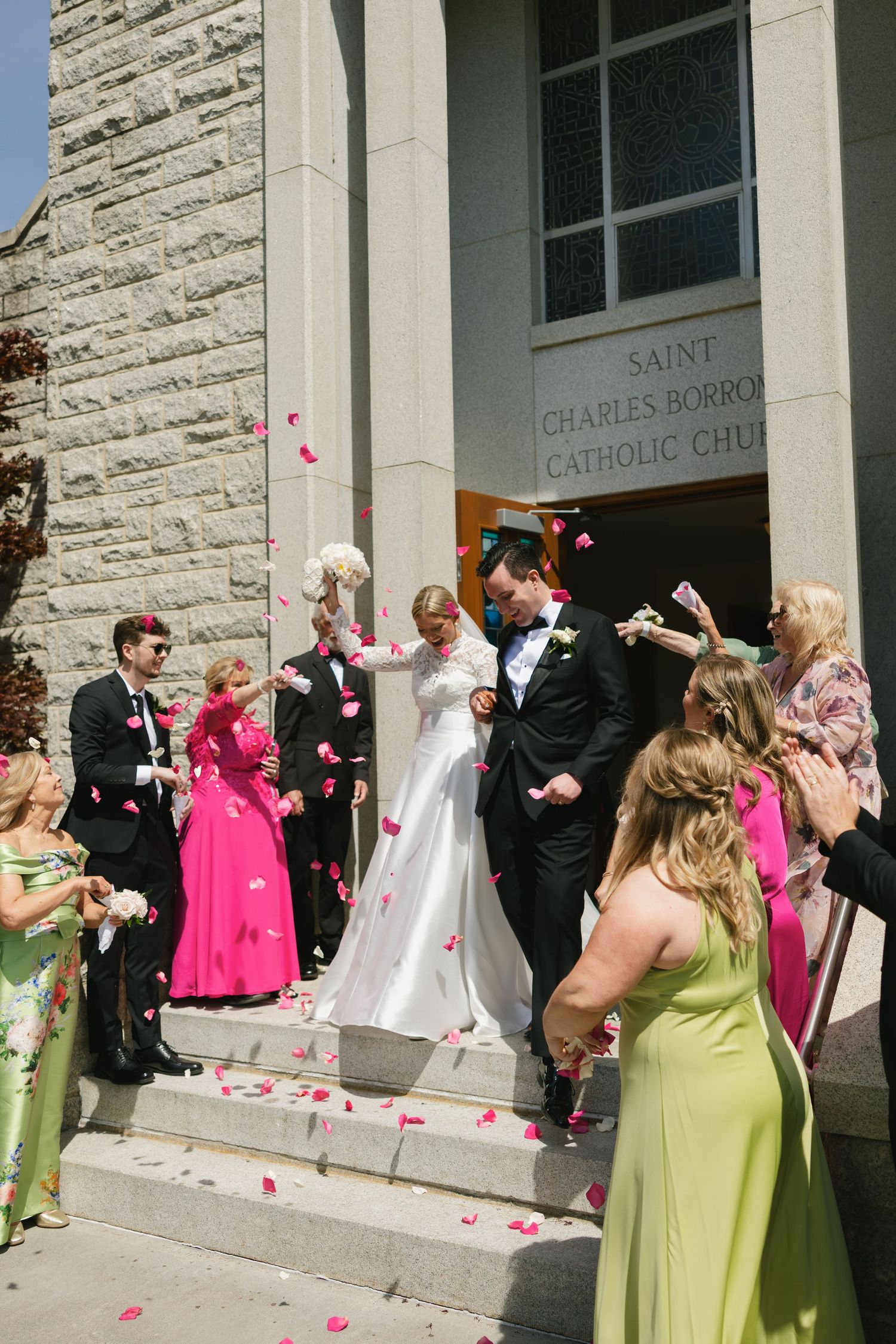Bride and groom exit Saint Charles Borromeo Catholic Church as guests toss pink rose petals in celebration.