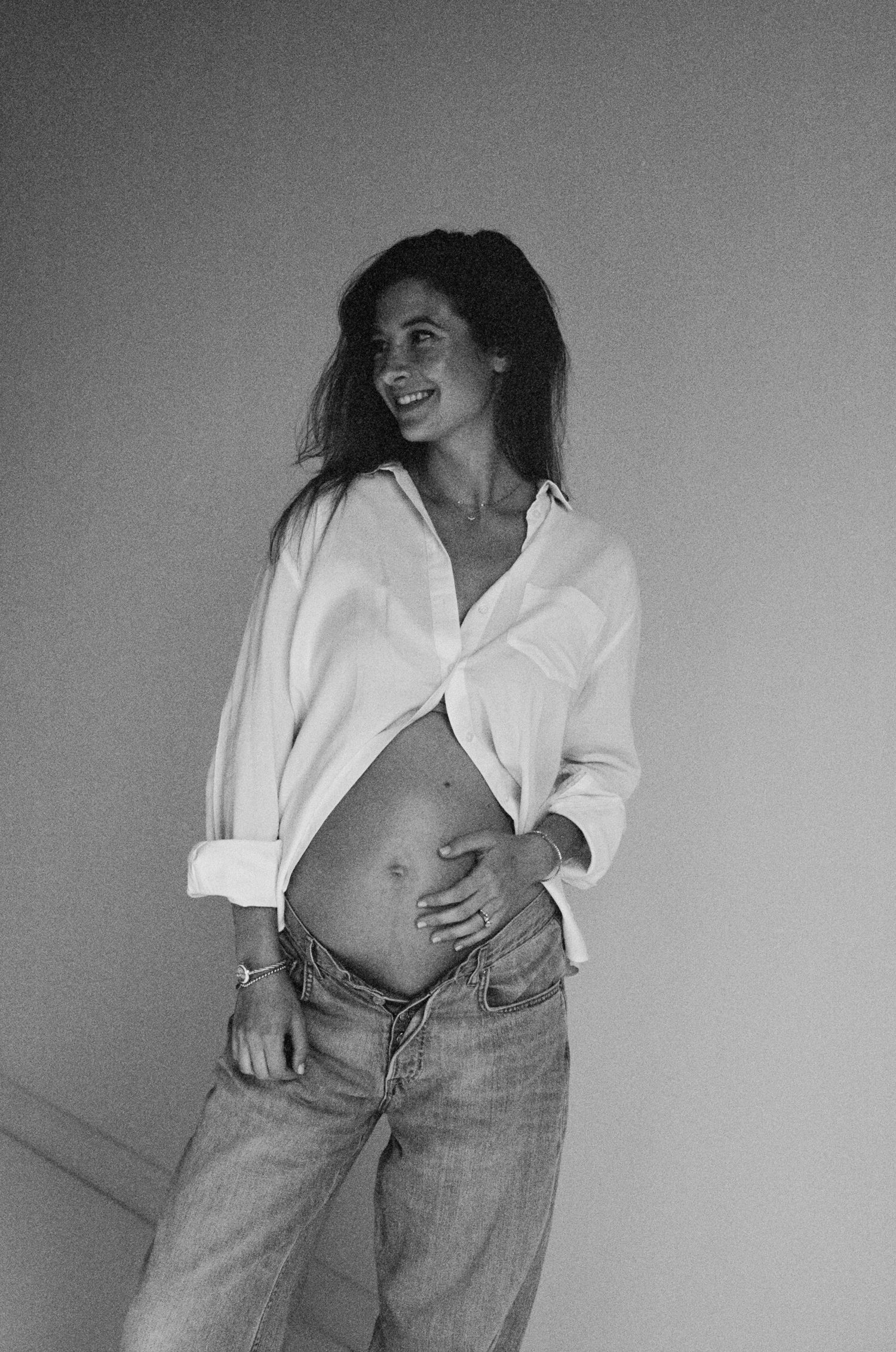 Black and white photo of a smiling pregnant woman in an open white shirt and jeans, cradling her baby bump.