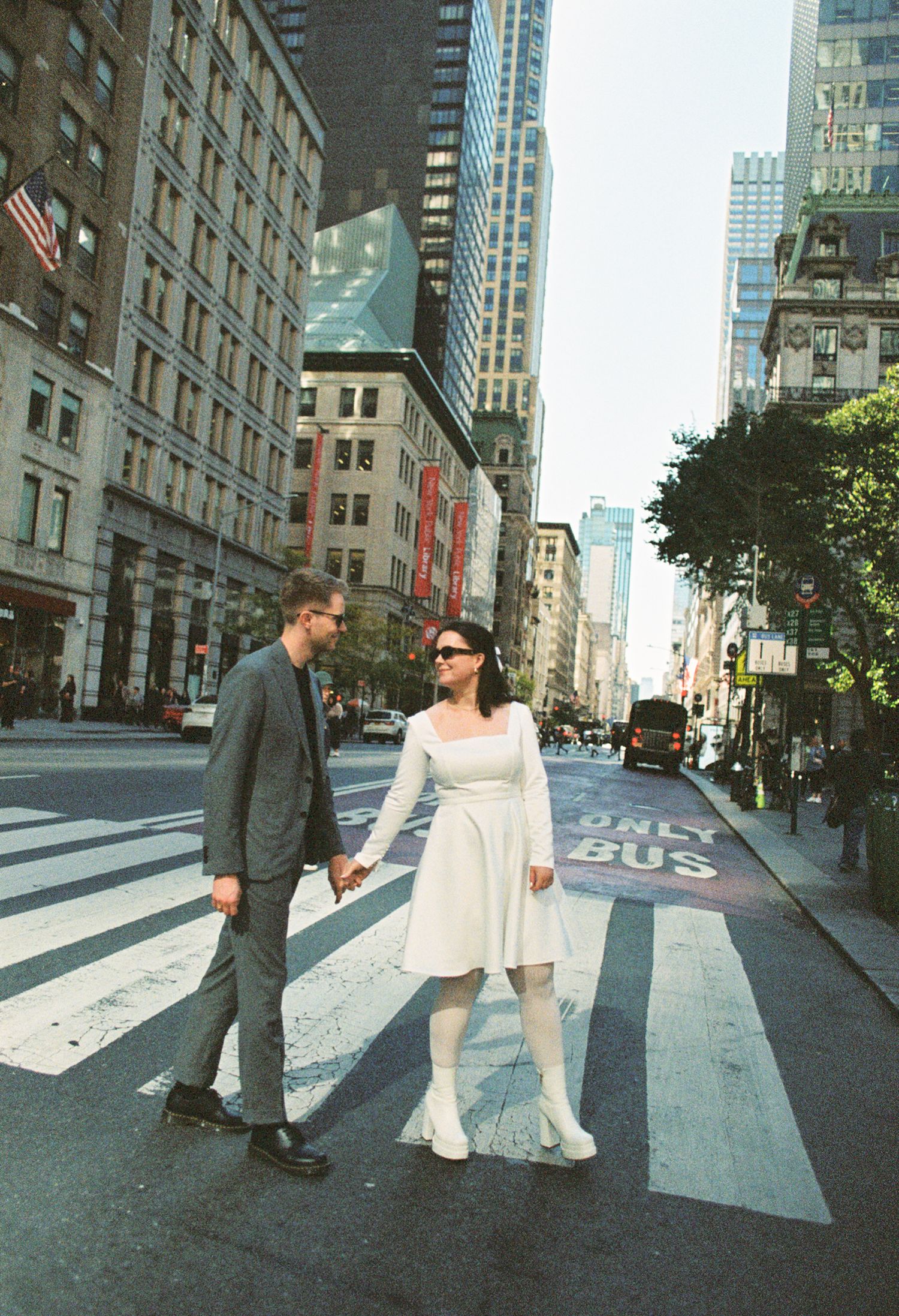 Couple holding hands crossing a zebra crossing on a busy New York City street, woman in white dress, man in gray suit.