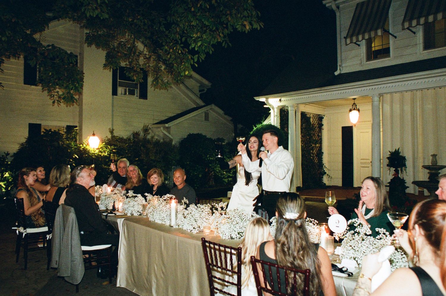 Wedding guests seated at a long candlelit table watch a bride and groom stand and toast outdoors at night beside a white historic house.
