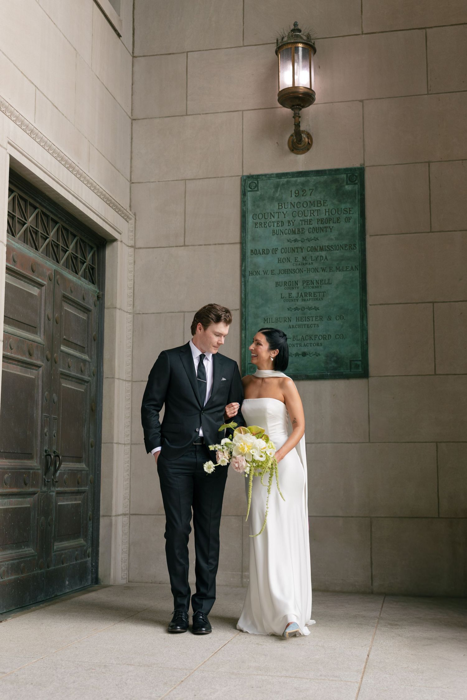 Bride in a white strapless gown holding a bouquet stands beside a groom in a black suit outside a stone building with a bronze plaque.