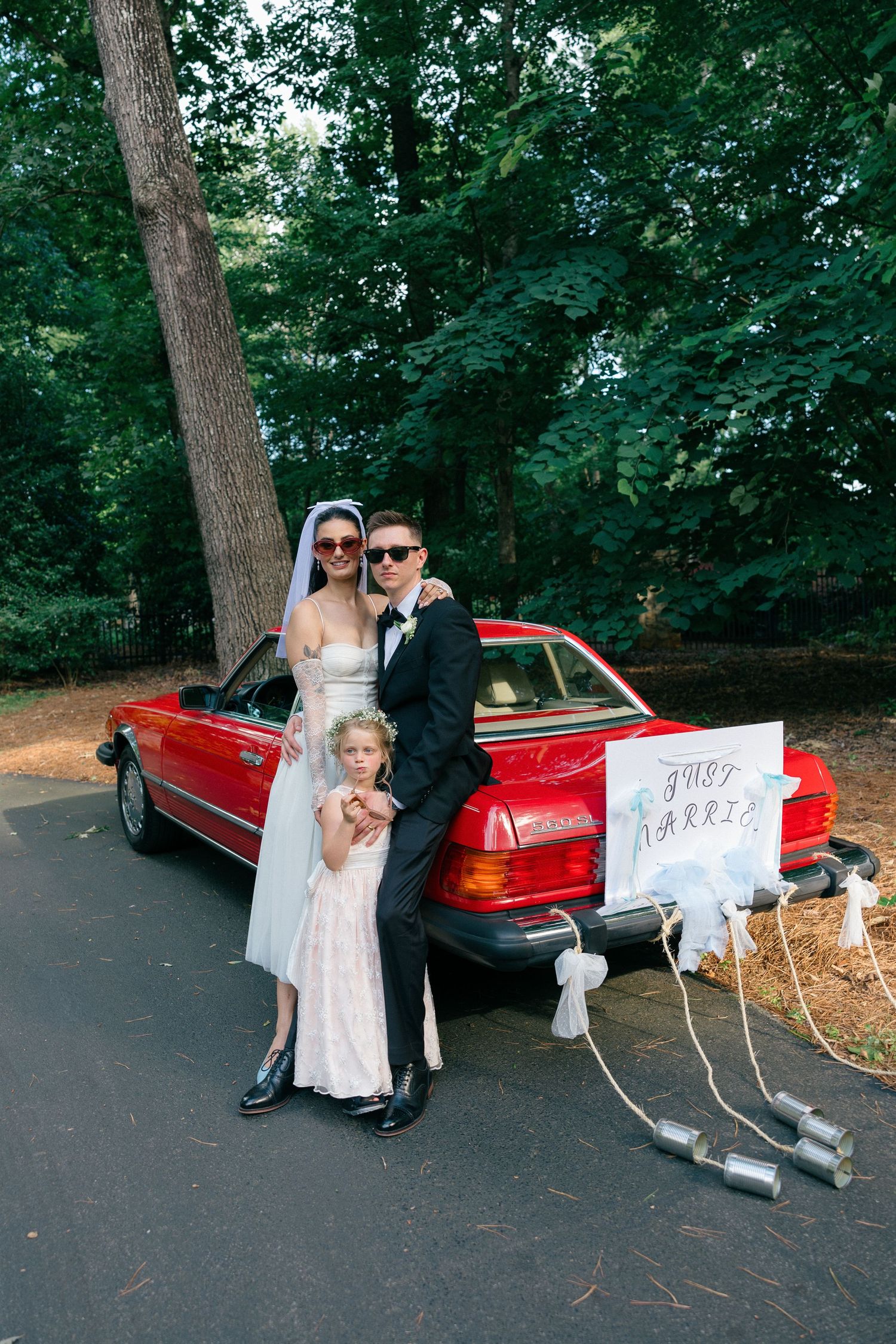 Bride and groom in sunglasses pose with a young girl beside a red convertible with a just married sign and tin cans tied to the bumper.
