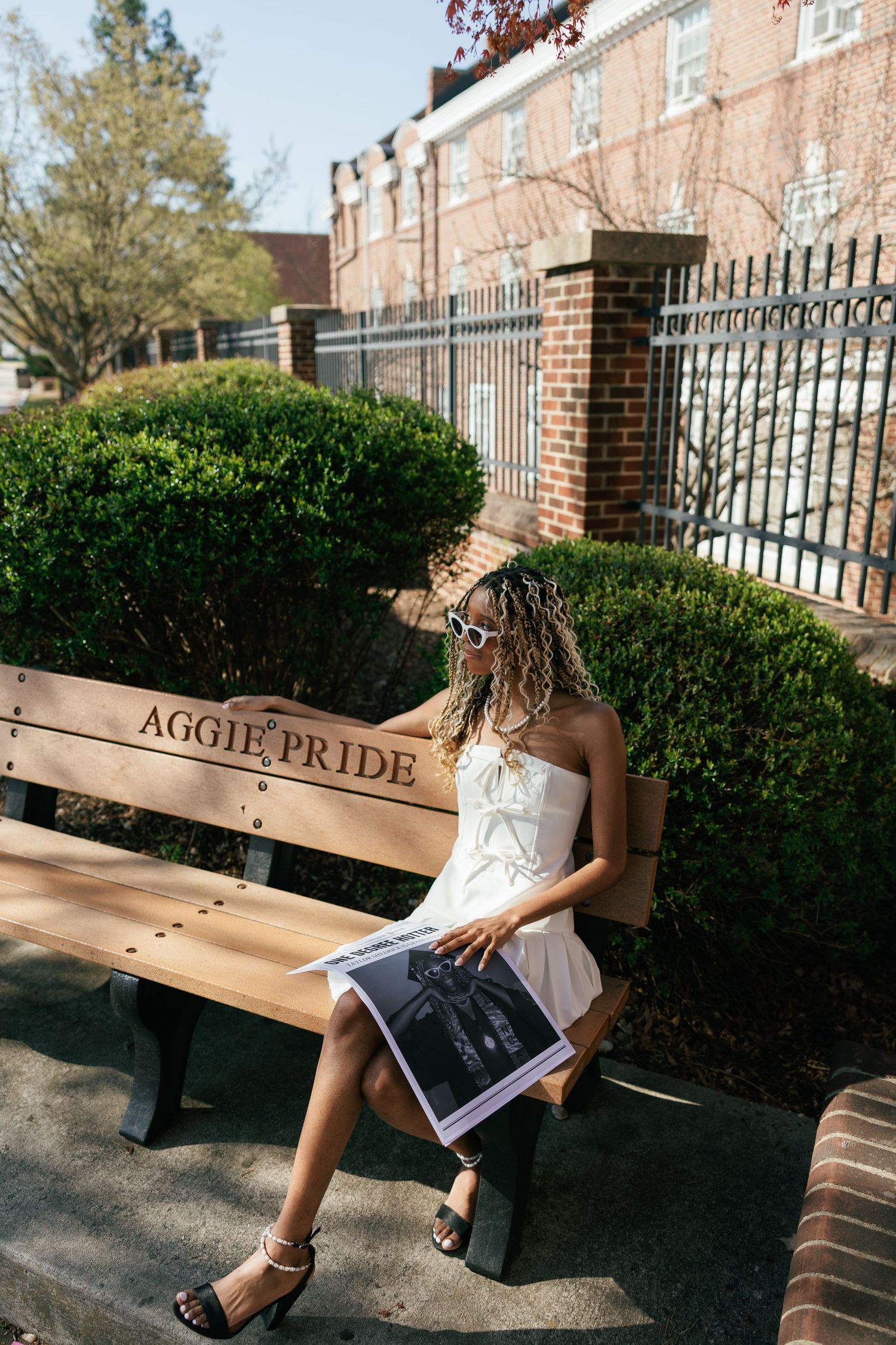 Someone in a white dress sits on an Aggie Pride bench near a brick building with manicured hedges and iron fencing.