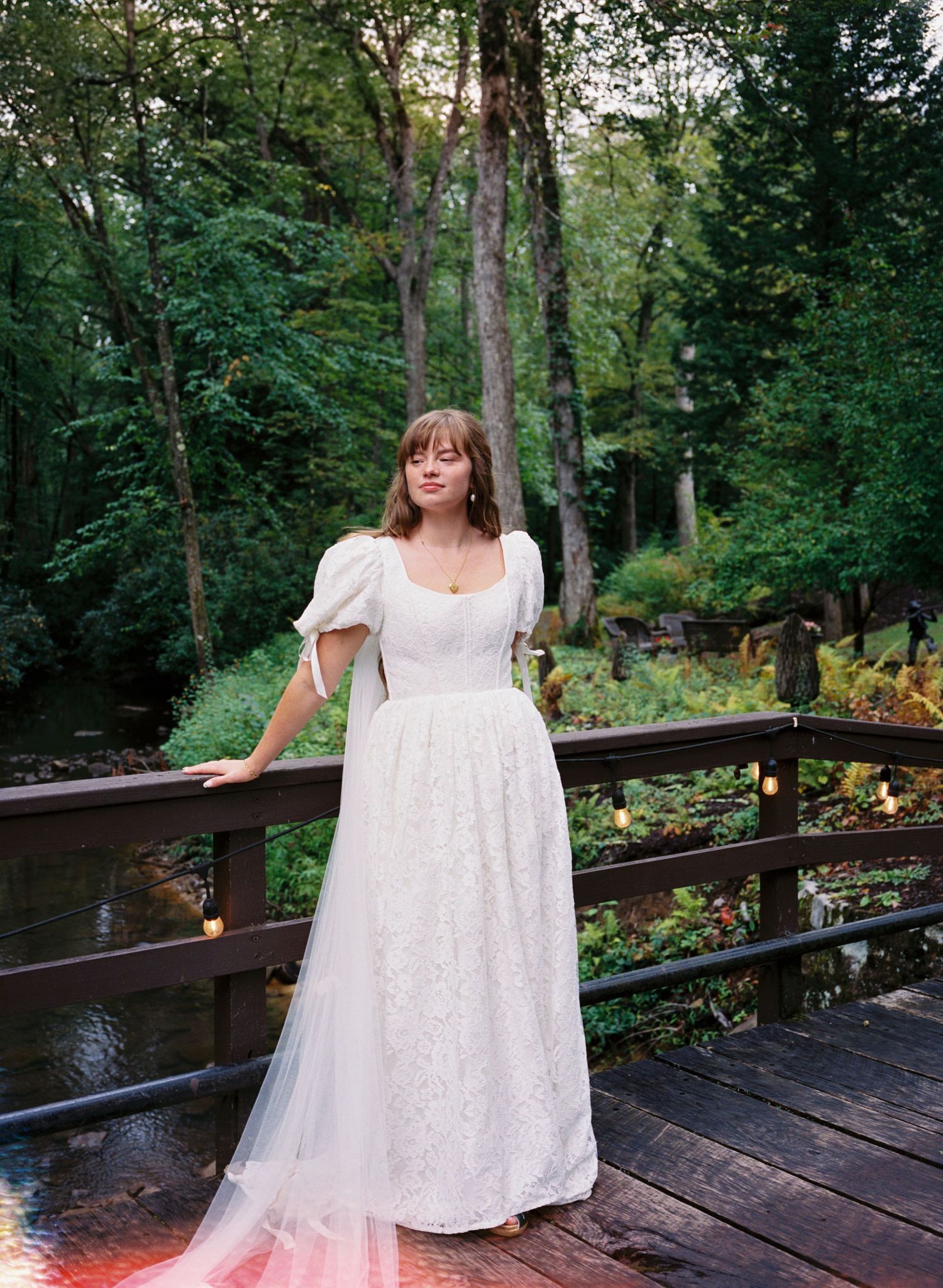 A person in a flowing white lace dress poses gracefully on a wooden deck surrounded by lush green forest scenery.
