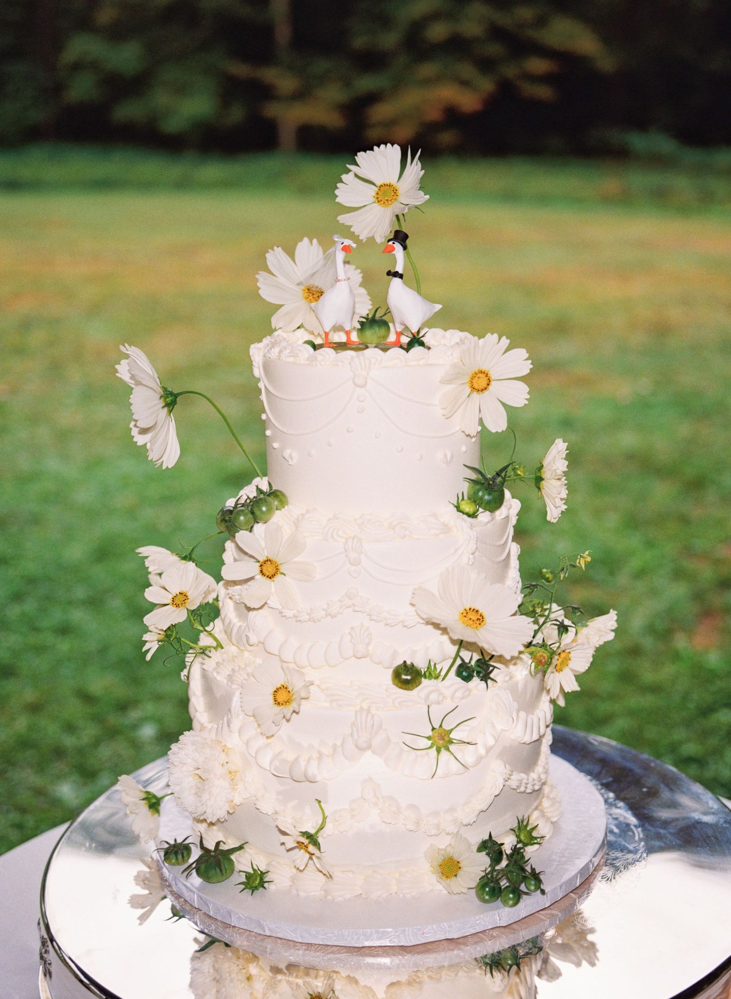 A white tiered wedding cake decorated with fresh white daisies and green ivy on a silver platter outdoors.