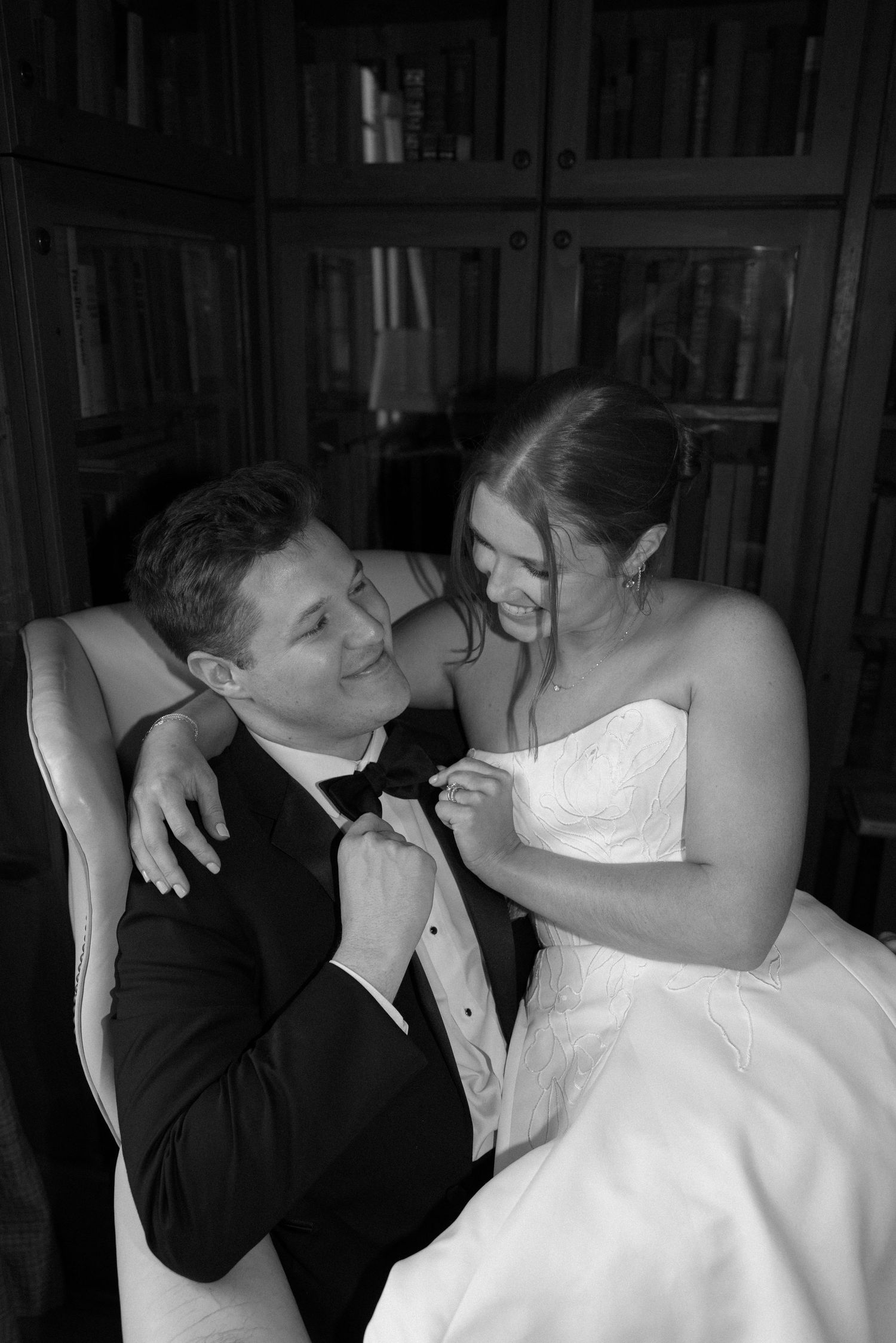 Black and white photo of a smiling bride and groom in formal wedding attire sitting together in a chair near a bookshelf.
