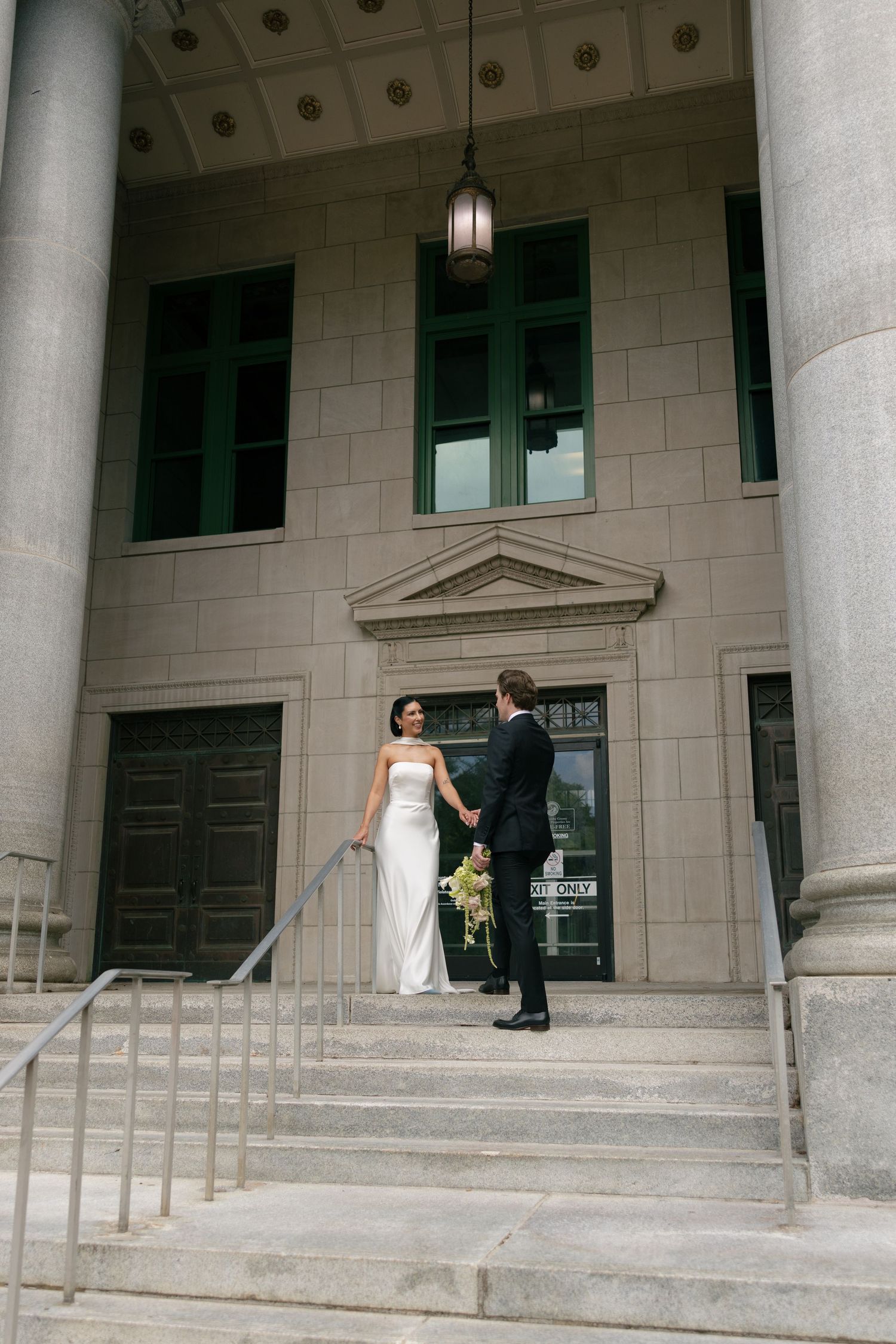 Bride in a white gown and groom in a dark suit hold hands on the steps of a stone courthouse with tall columns and green-framed windows.