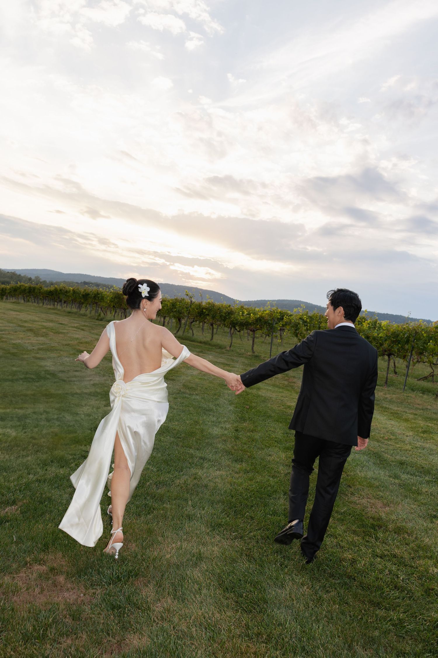 A couple holding hands and walking through a vineyard at sunset with mountains in the background.