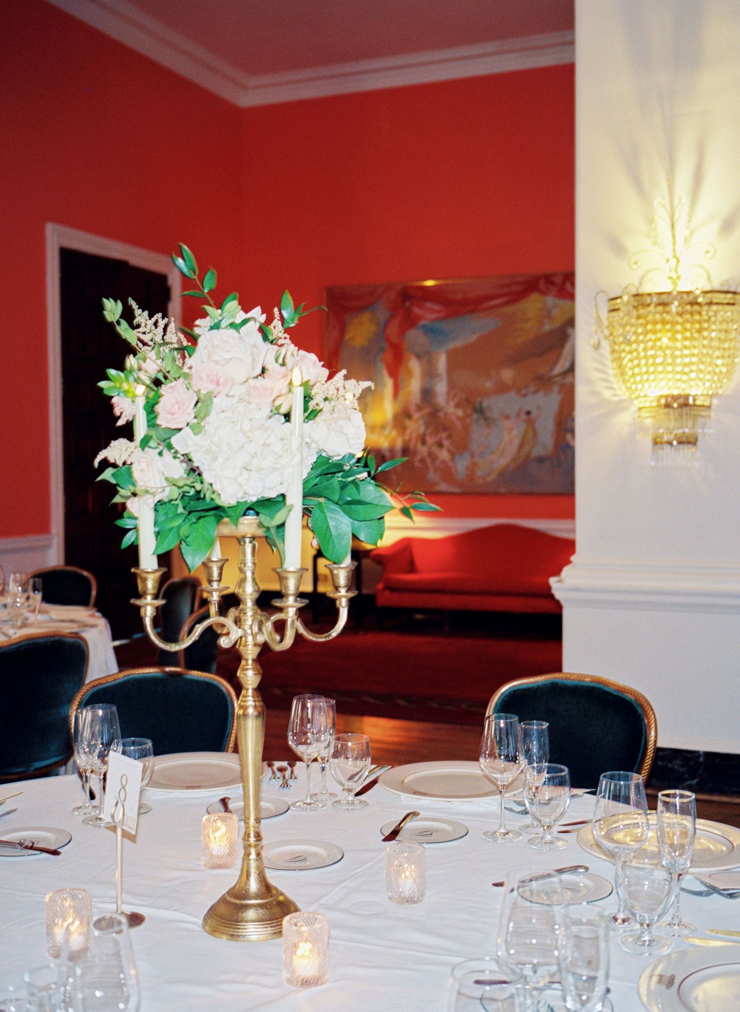 Elegant wedding reception with gold candelabra centerpiece, white florals, crystal sconce, and red walls.