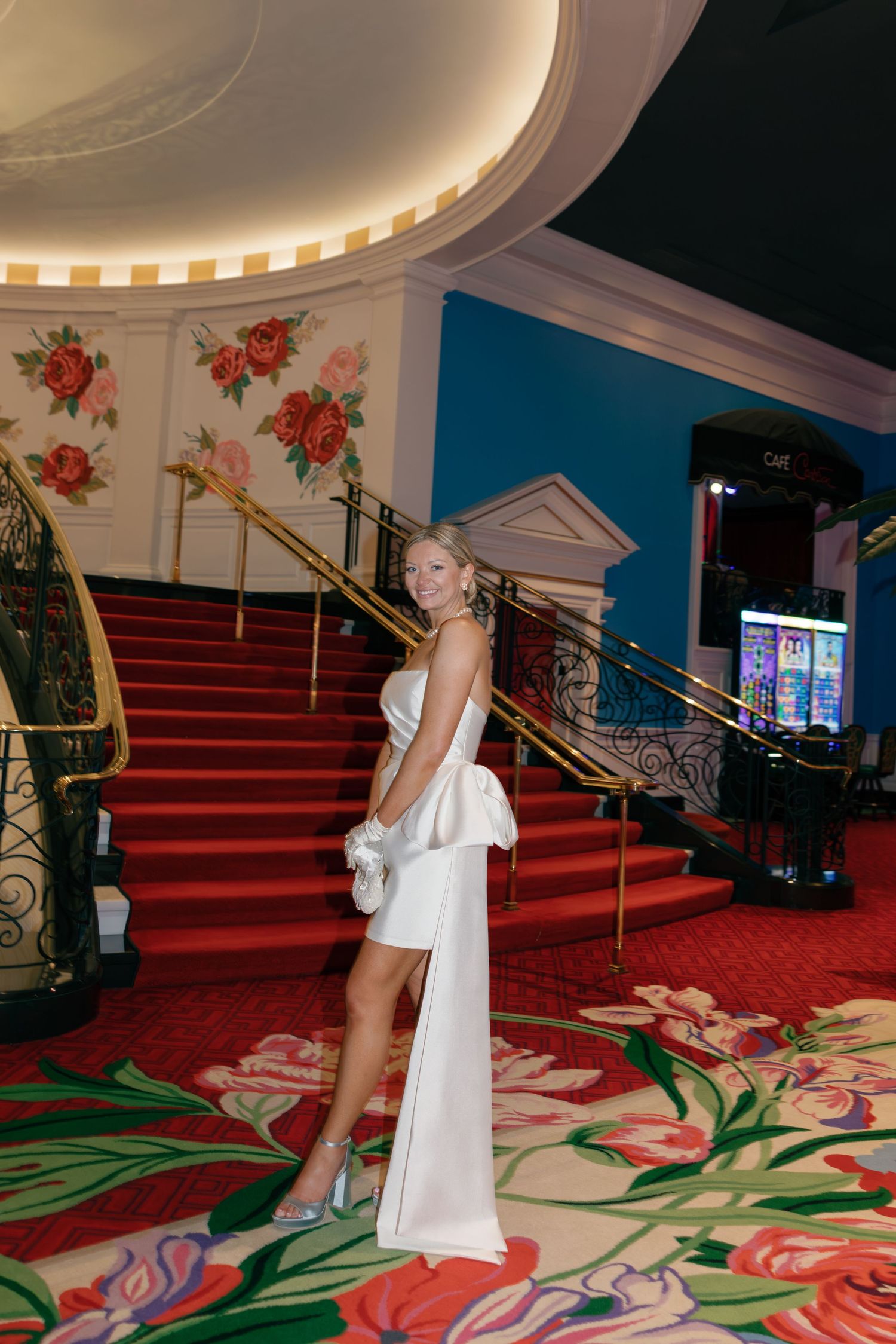 Elegant woman in white strapless mini dress poses on red carpeted staircase in ornate venue with floral decor.