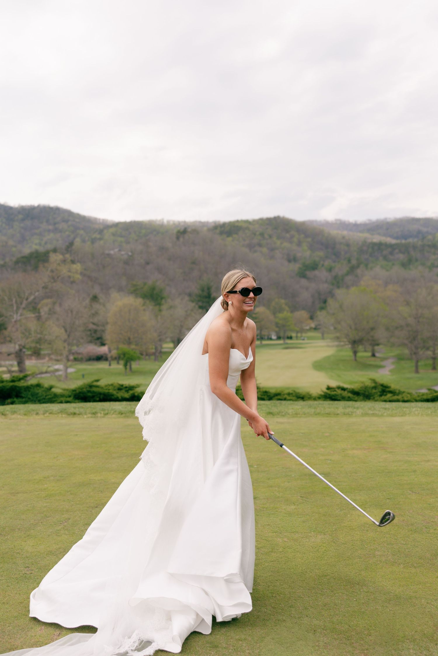 Bride in white gown and sunglasses playing golf on a lush green course with mountain views.