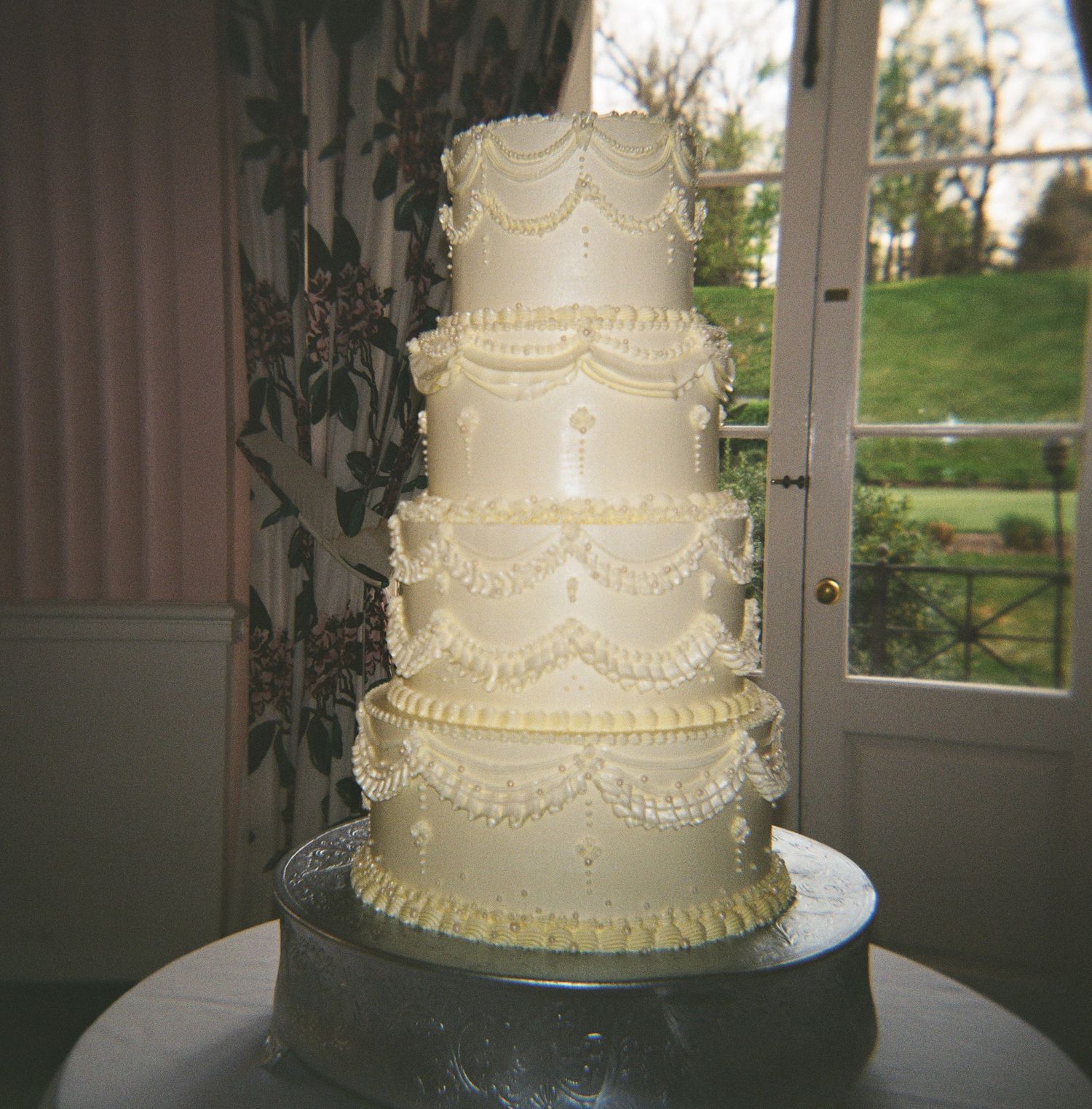 Elegant five-tier white wedding cake with intricate piped lace details and pearl accents on a silver stand.