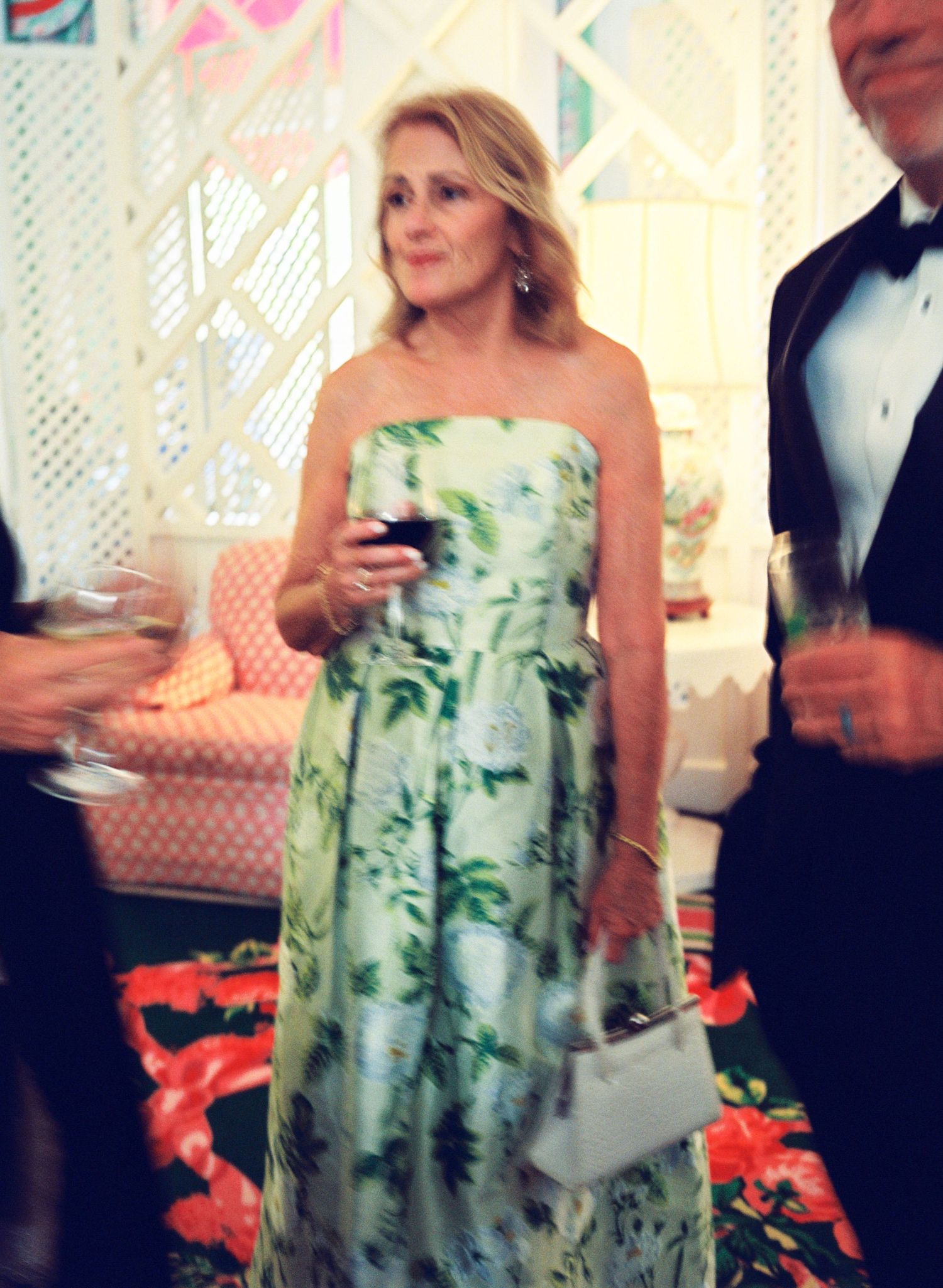 Woman in strapless green floral gown holding wine glass at elegant indoor social event.